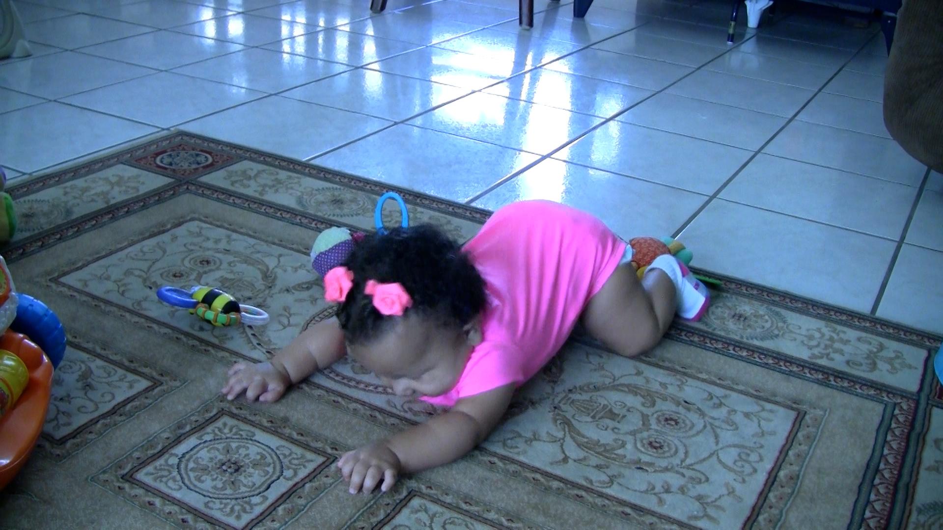 Baby girl wearing pink onesie and pink rose hair clips pushing backward with her arms while on her knees.