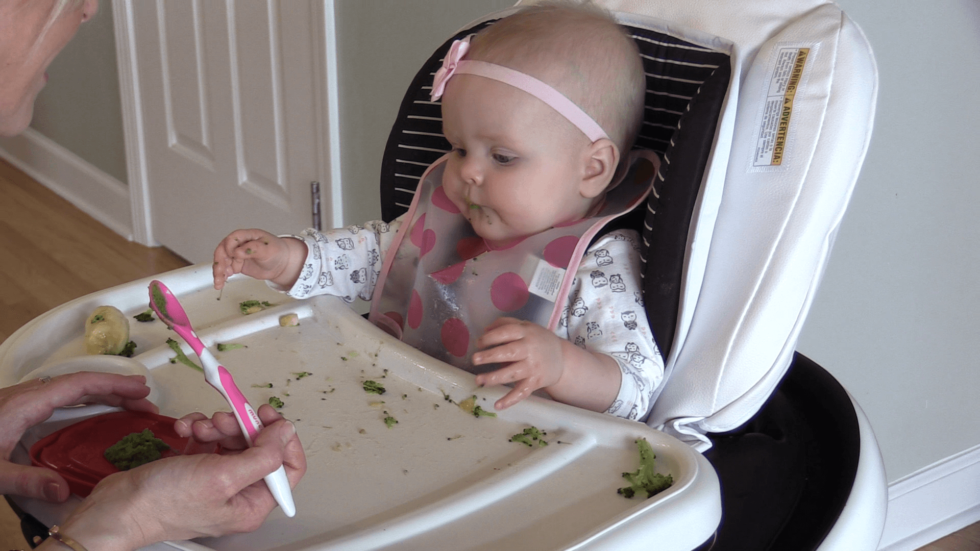 Baby girl with pink bow headband and pink polka dot bib sitting in high chair with broccoli mushed all over it. Mom holds pink spoon with green puree in front of baby.