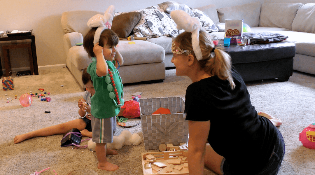 Little boy and woman playing dress up with bunny ears while sitting in a living room scattered with toys.