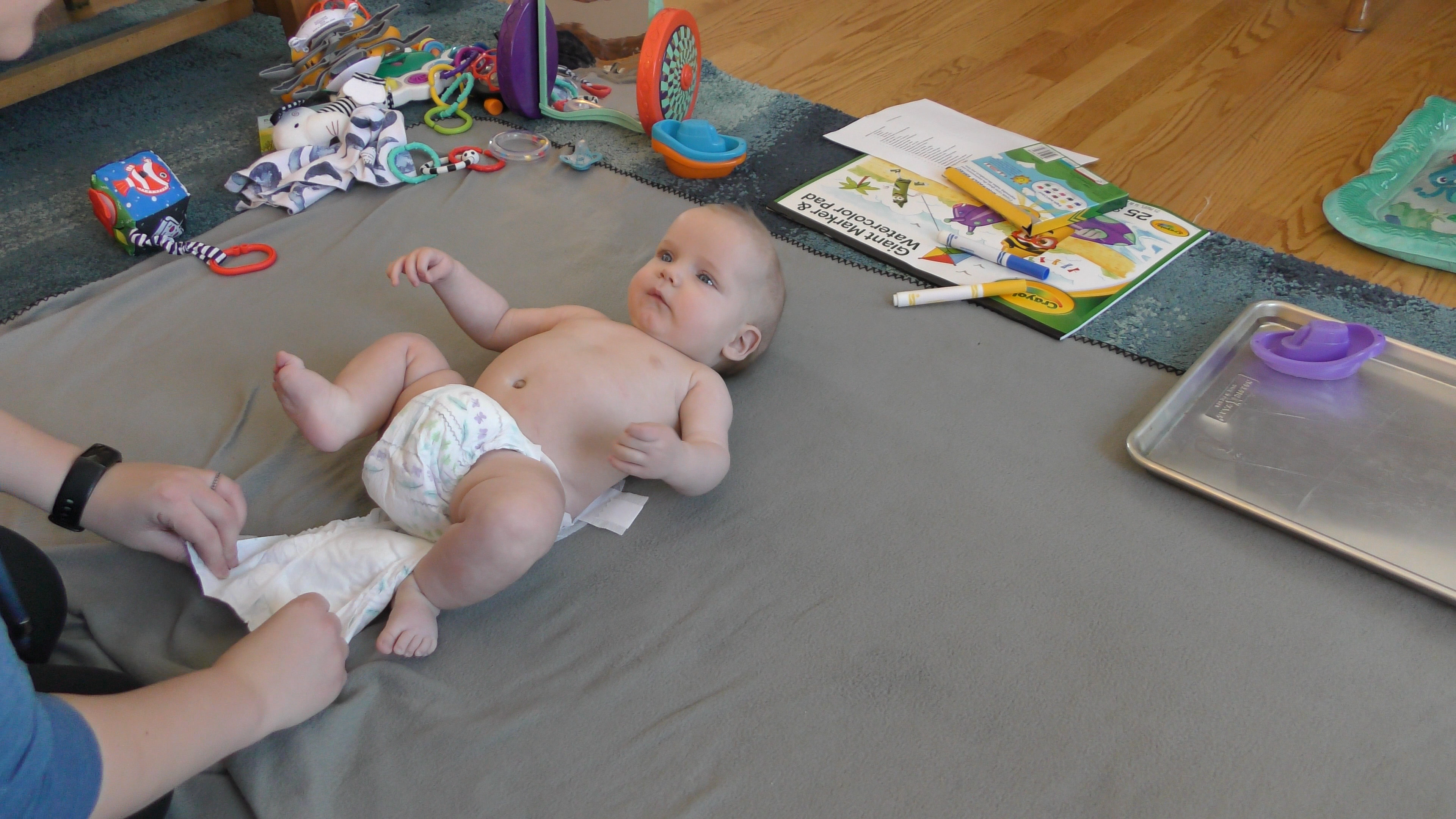 Baby boy having his diaper changed on a blanket surrounded by toys on a living room floor. A clean diaper is positioned under the dirty, unfastened diaper.