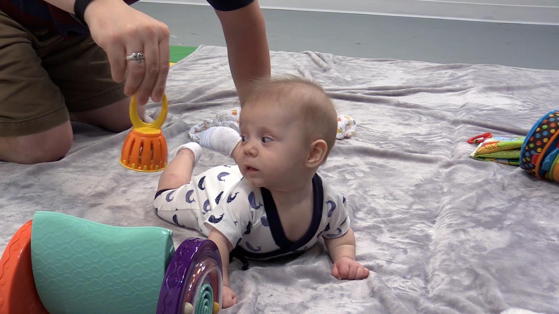 Baby boy lying tummy-down on a soft gray blanket. He is propped up on his forearms and turning his head to the side to look at a toy being dangled in the air.