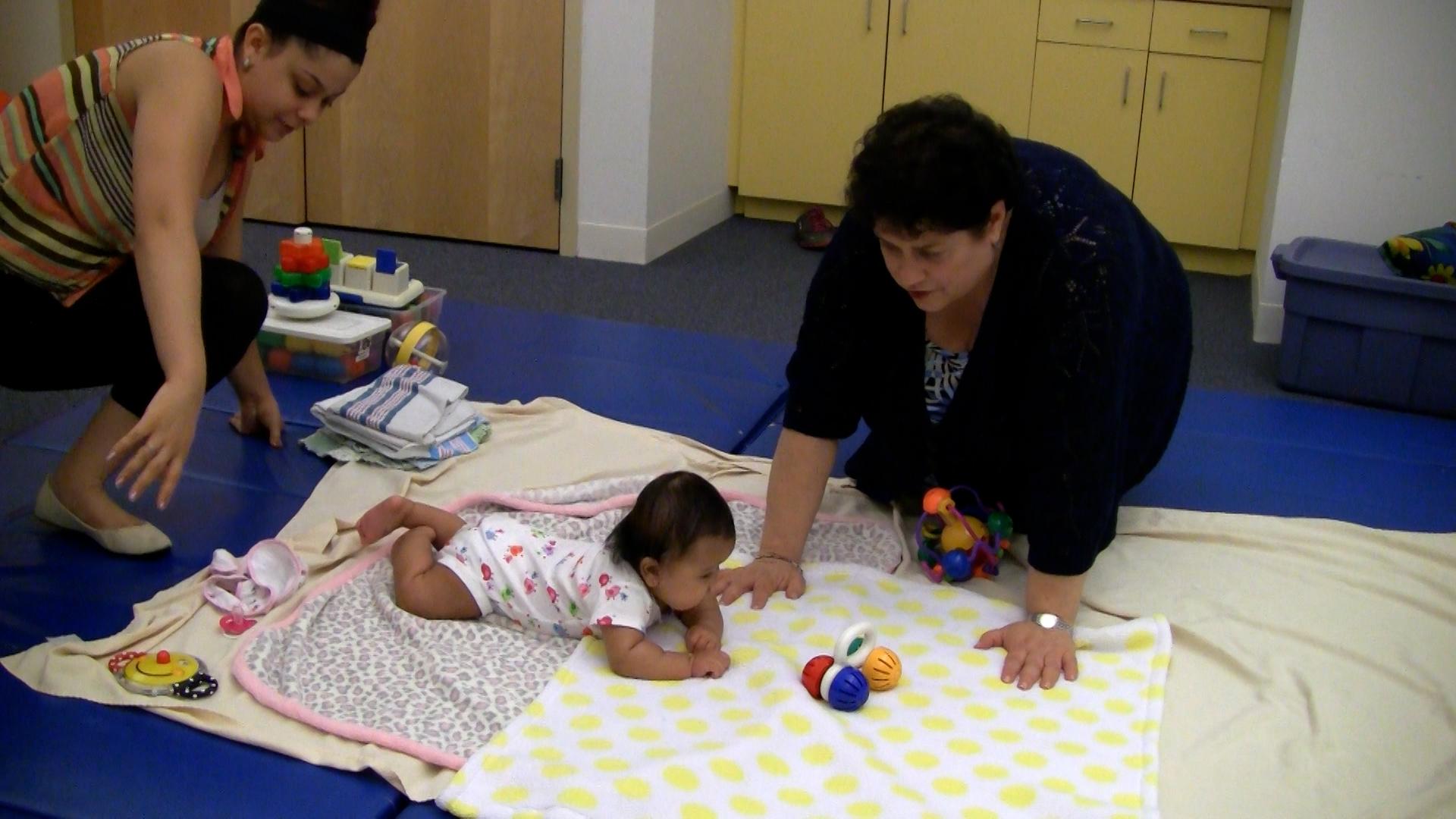 Two caregivers sit with a baby girl, who is doing Tummy Time on top of patterned blankets. They are in a playroom surrounded by colorful baby toys.