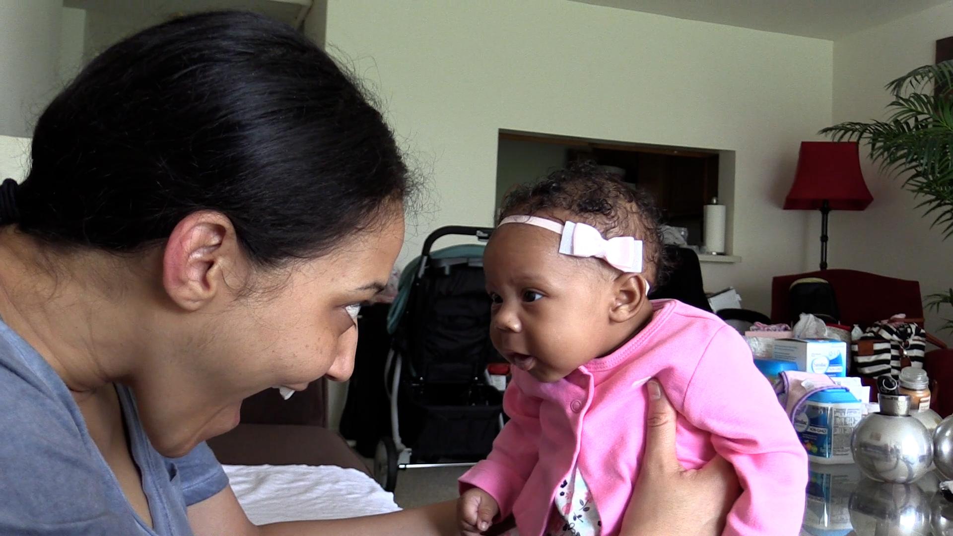 Close-up side view of a smiling mom and newborn baby girl making eye contact. The baby girl is dressed in a pink cardigan and a light pink hair bow.