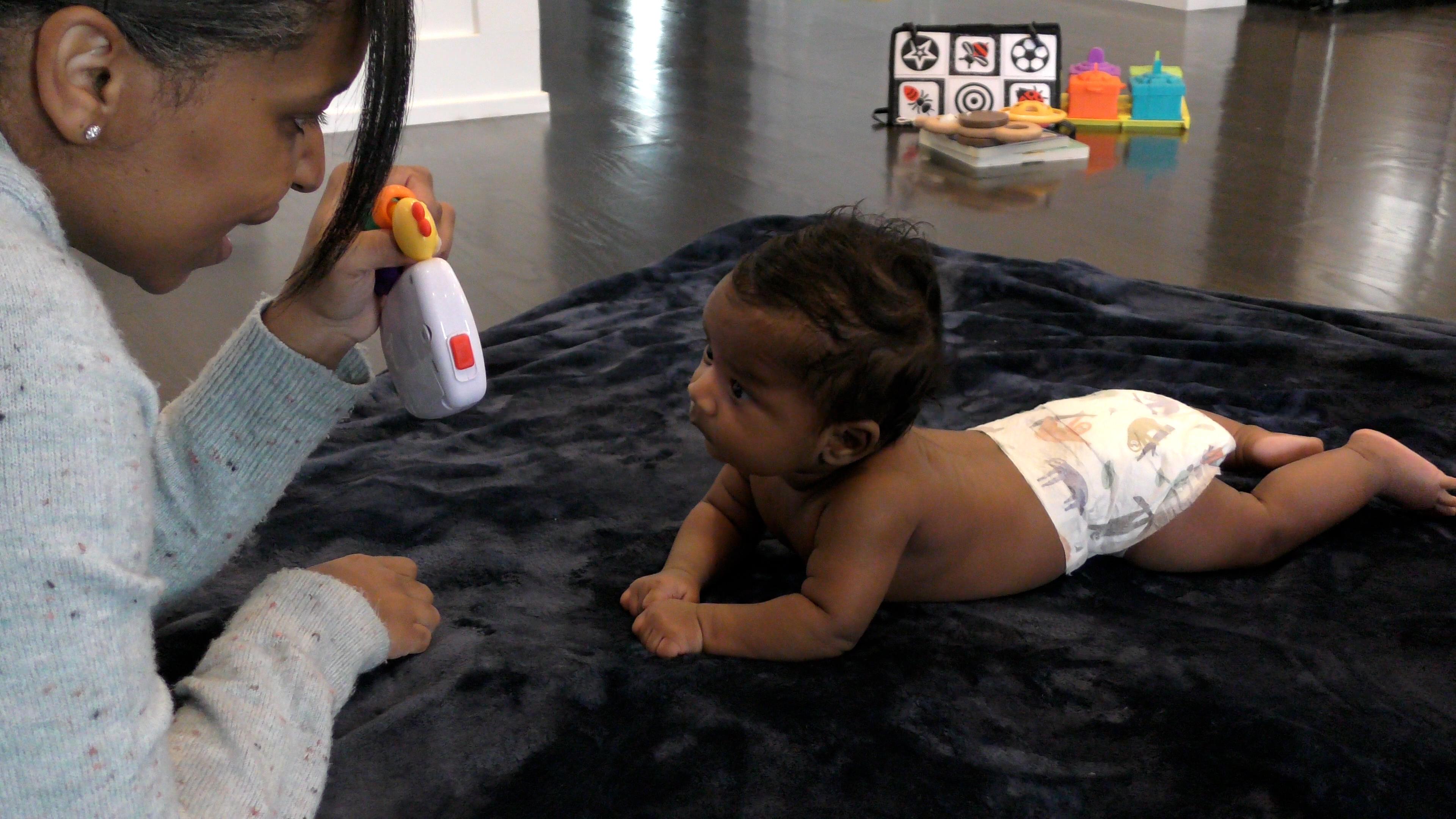 Mom and infant boy lying on their stomachs, face-to-face on a navy blue blanket. The baby holds his head up to look at a toy the mother is holding. A small pile of baby toys is seen in the background.