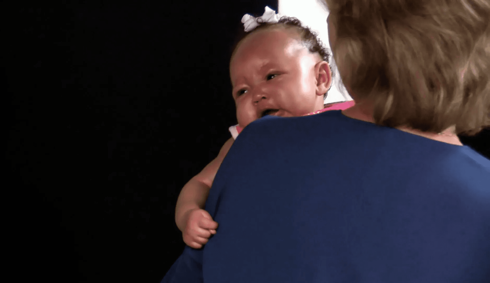  Baby girl with a small white bow on her head crying while being held over a woman’s shoulder.