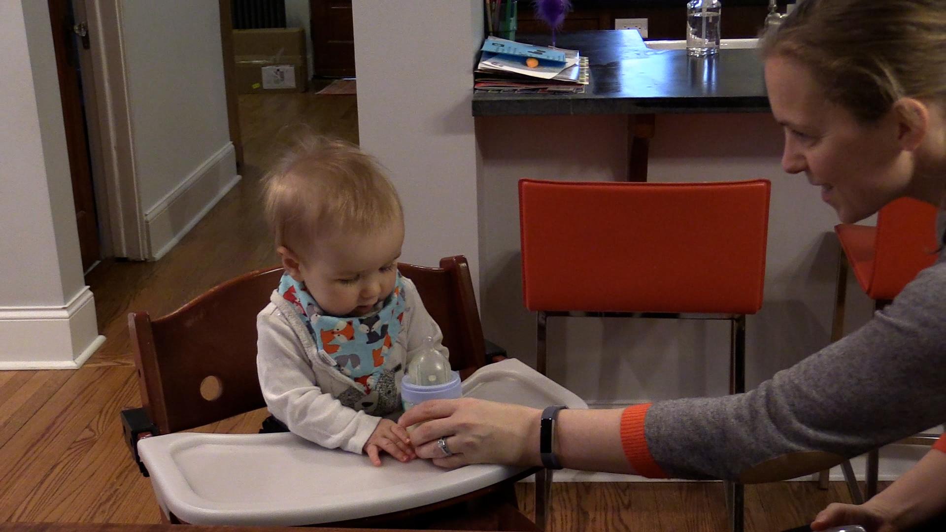 Mom handing baby boy a bottle of milk while he sits in a high chair.