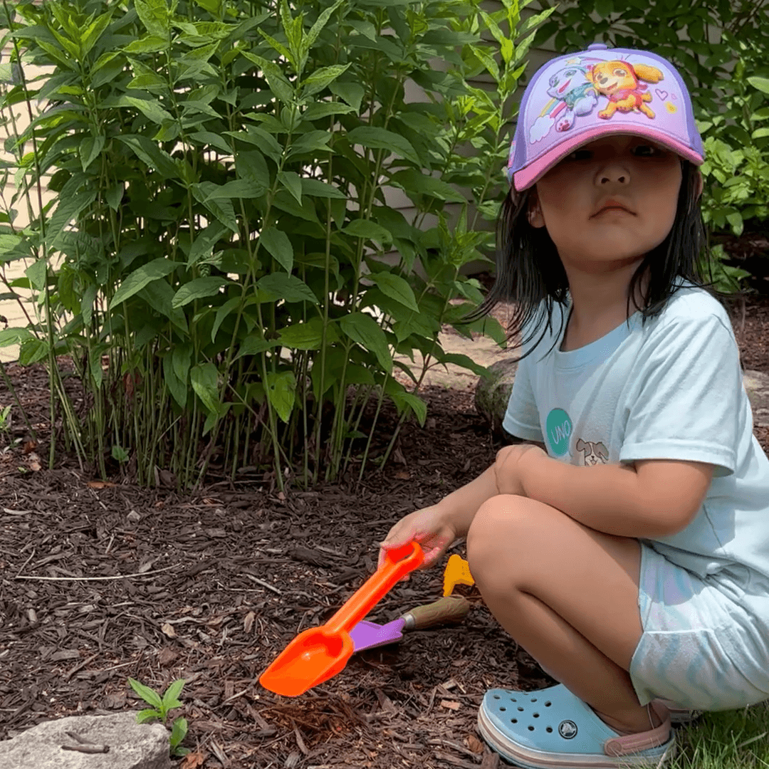 Toddler girl dressed in a hat and light clothing plays in the dirt with toy shovels.