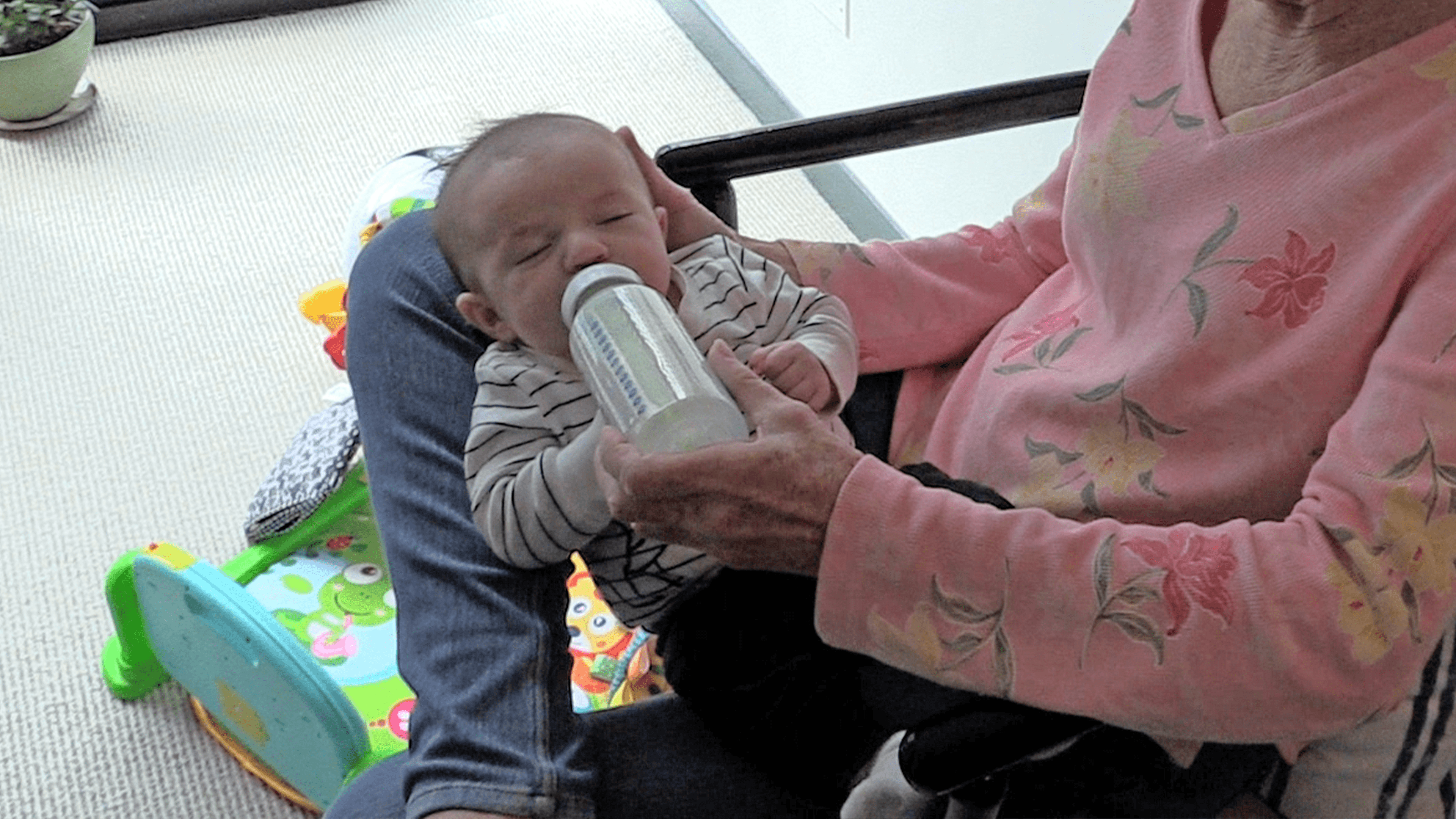  A grandmother in a pink floral shirt bottle feeds a baby boy, who is in a black-and-white striped shirt and is lying across his grandmother’s lap. A brightly-colored, animal-themed baby gym can be seen in the background.