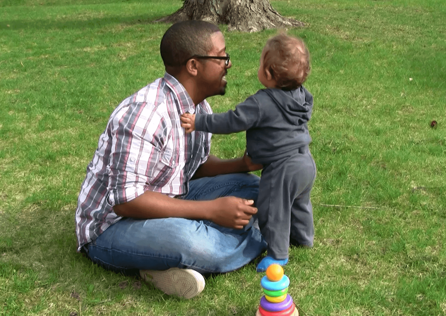 Dad sits criss-cross on grass lawn while baby boy stands with outstretched hand resting on dad’s chest for support.