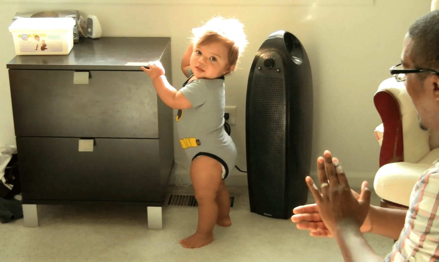 Baby boy stands independently with hand resting on short bedside table while dad claps encouragingly.