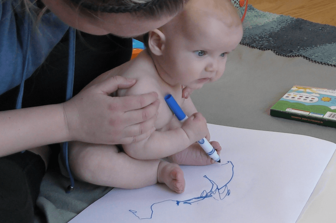 Baby boy making marks on paper with blue marker while sitting with support from his mother.