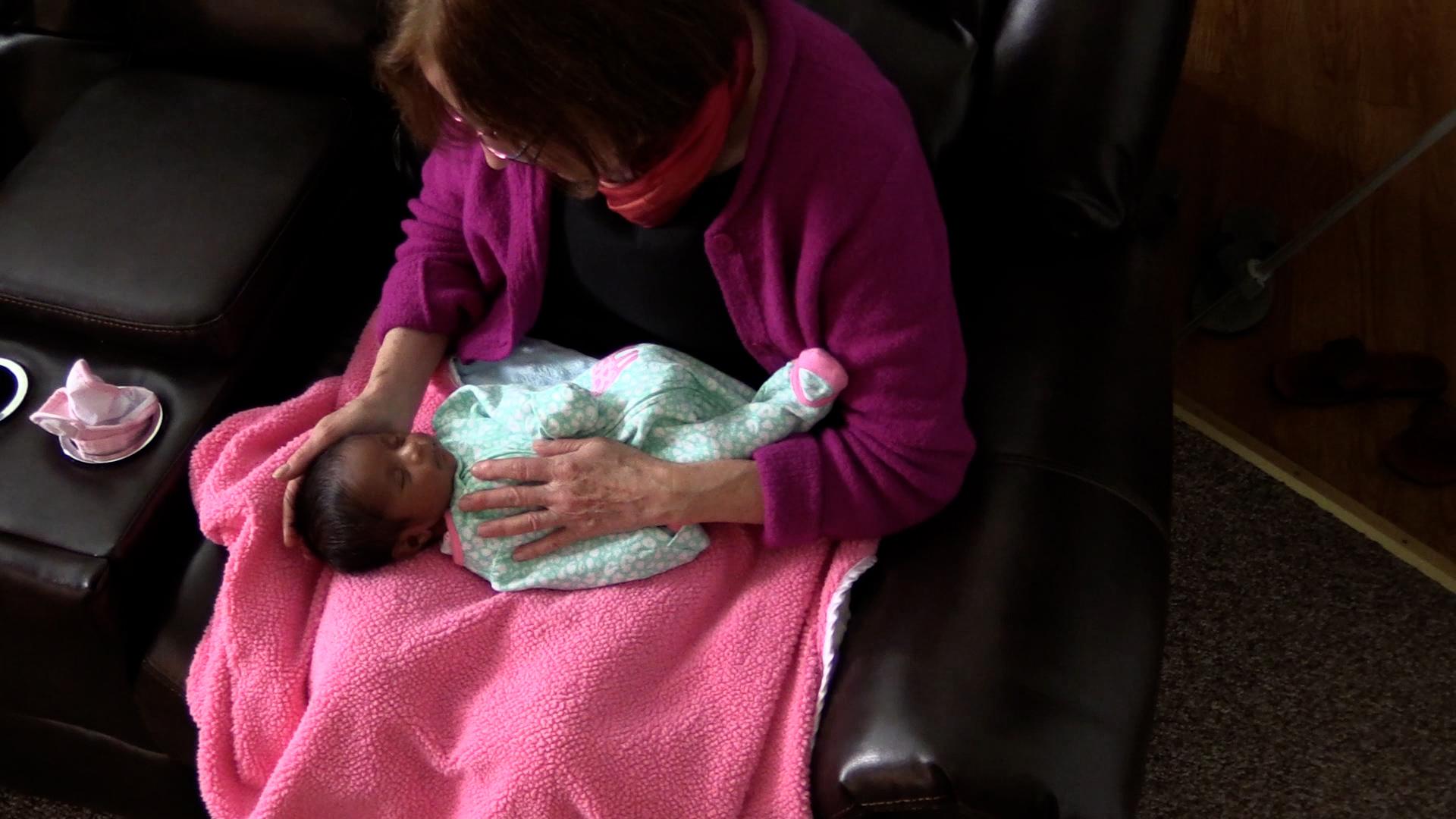 Older woman in a magenta cardigan sitting in a leather chair with a baby lying across her lap atop a hot pink blanket. The baby has a peaceful expression while the woman performs a calming baby massage.