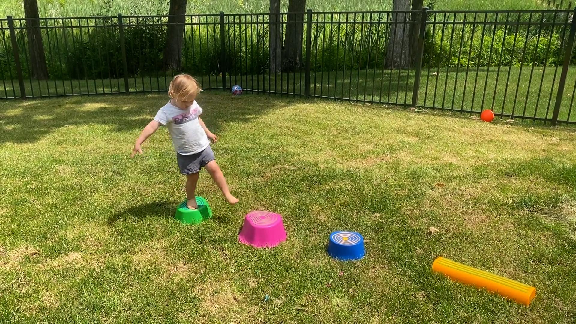In a backyard setting, a toddler girl is mid-step on an obstacle course made of stepping stones in various colors and shapes.
