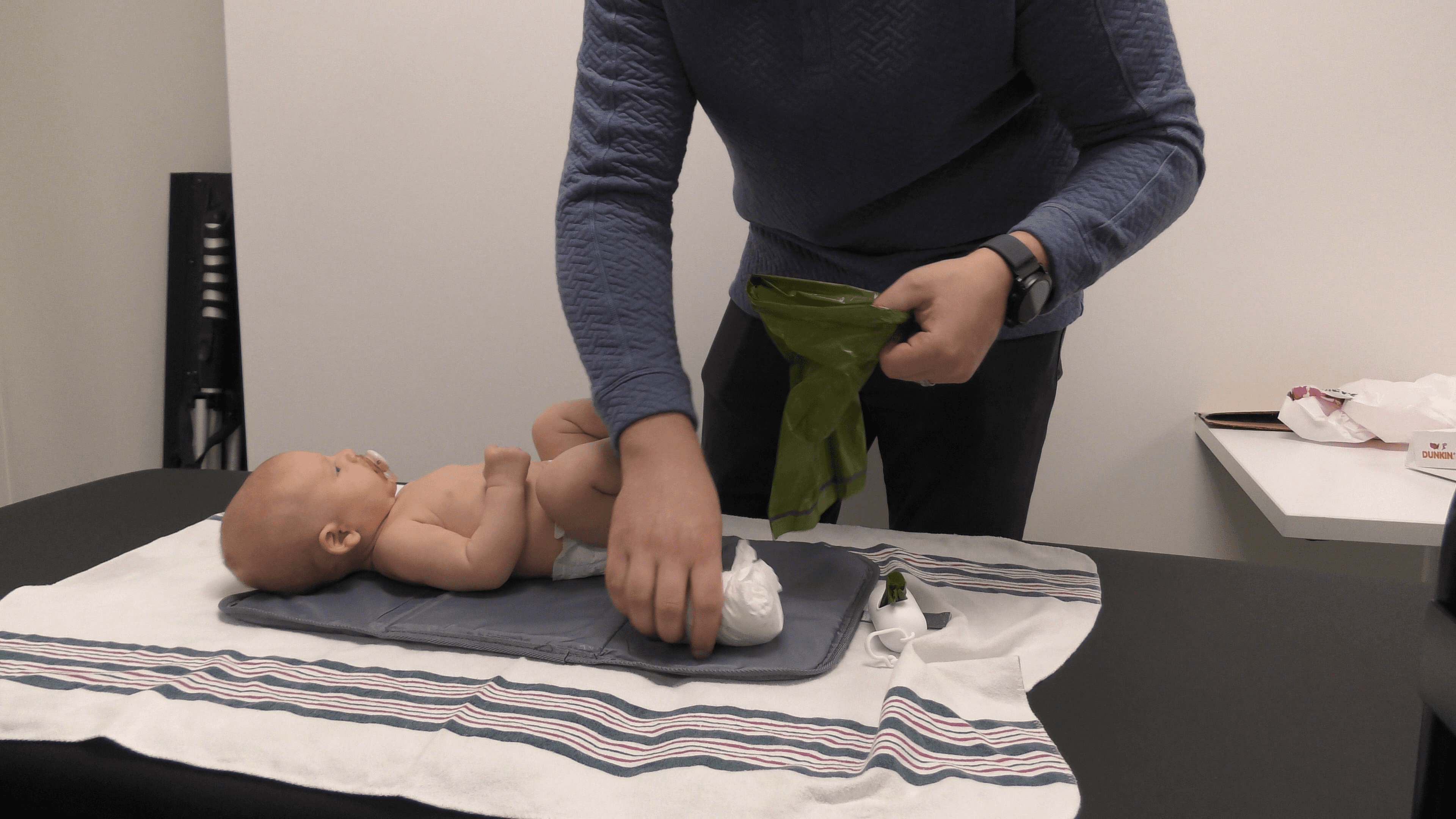 Baby boy lies on a table after a diaper change while his father puts his dirty diaper in a dog waste bag.