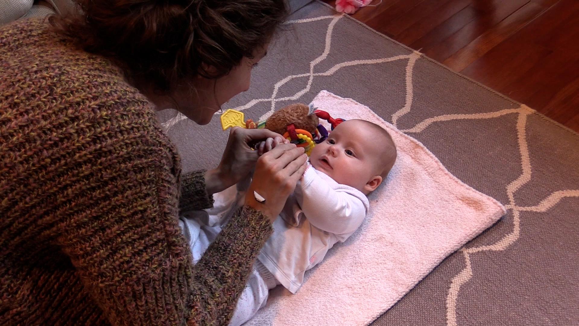Angled top-down view of a mom holding her baby’s hands together in midline while the baby lies on her back. The baby is on a pink blanket with a colorful toy near her head.
