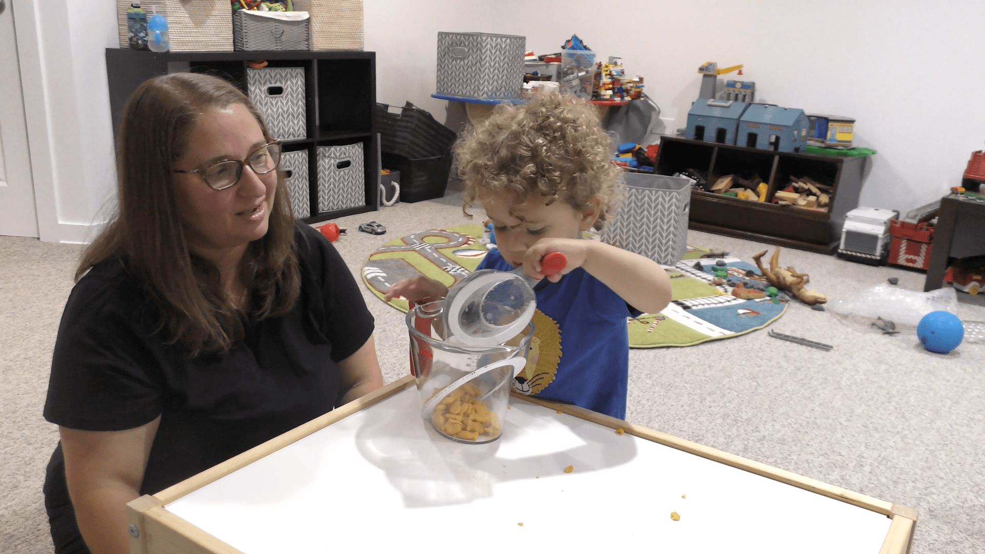 Curly-haired toddler boy pouring cheese crackers from a small measuring cup into a big measuring cup while standing at a kids’ table next to his mom.