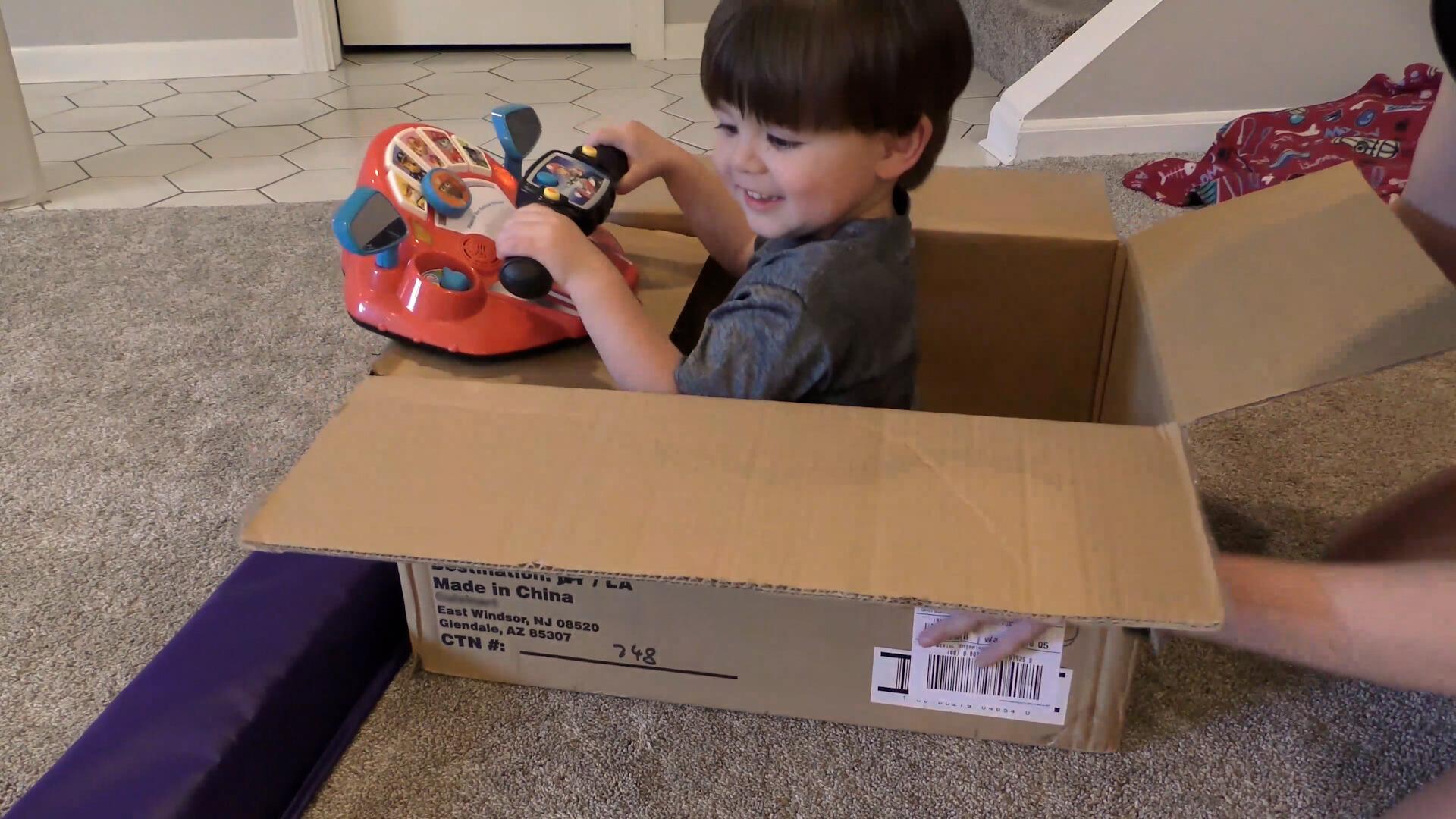 Toddler boy being pushed in cardboard box pretending to drive with a toy steering wheel.