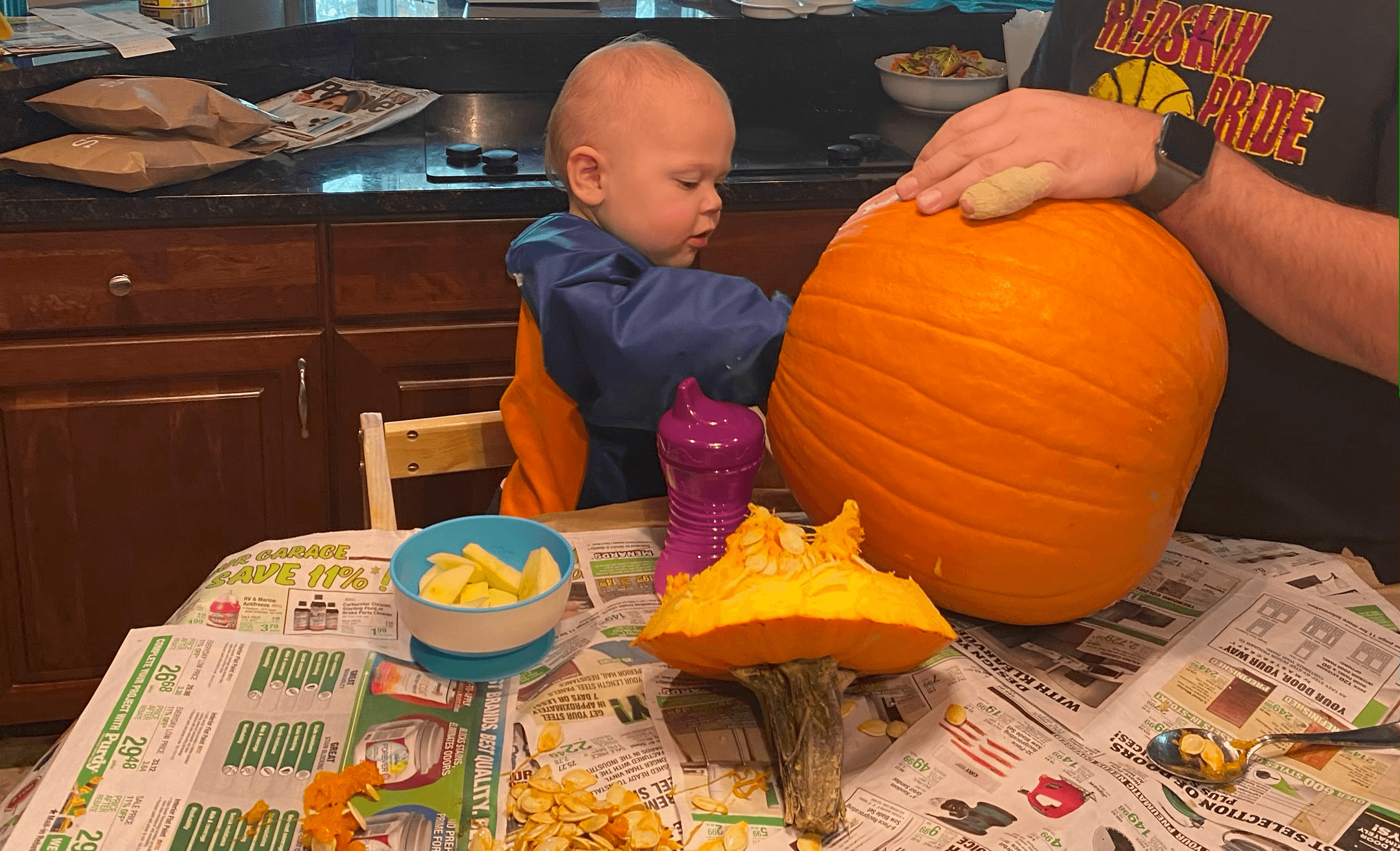 Toddler boy reaching into a round pumpkin held by his dad to scoop the guts onto the table covered with newspaper.