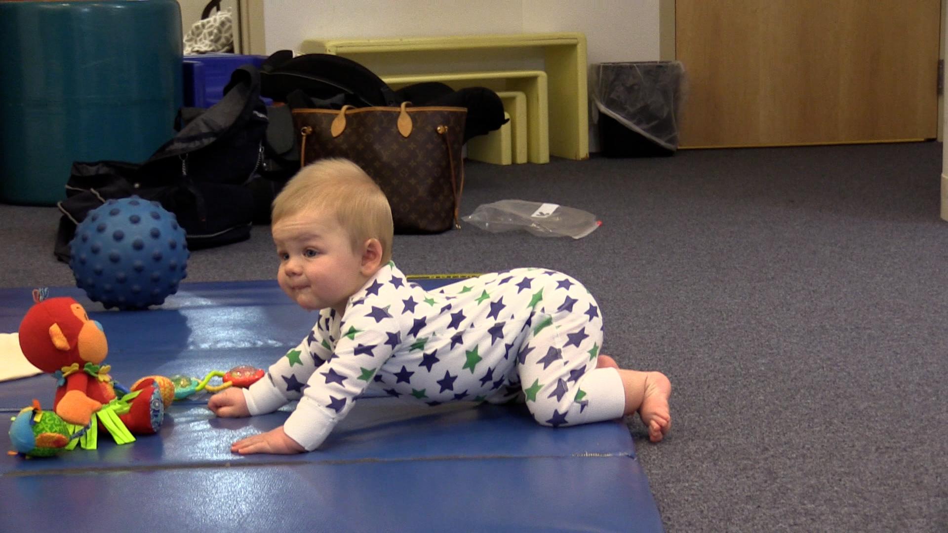 Baby boy in star-patterned jumpsuit scooting backward on a blue playmat with toys in front of him.