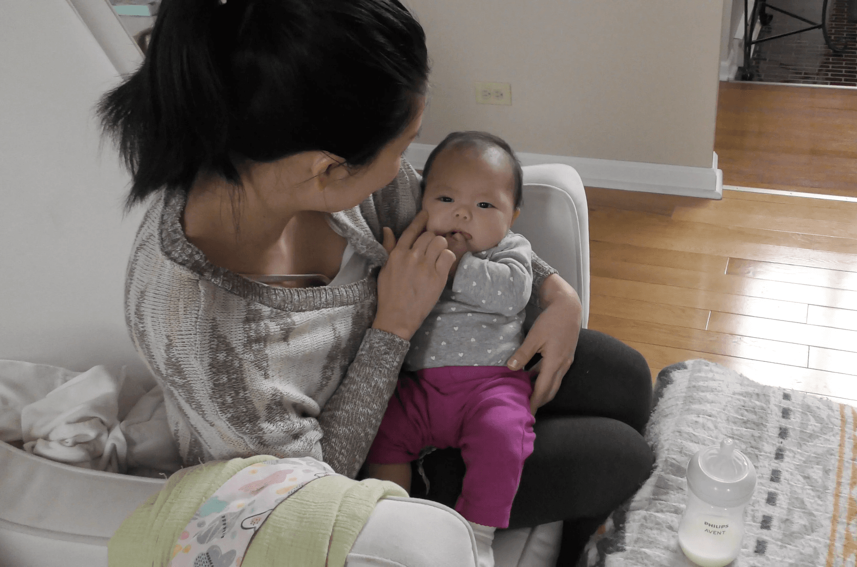 Mom, faced away, sits in an armchair while holding her baby in her lap and caressing her cheek. The baby, looking ahead, has one hand in her mouth. A milk bottle sits on a table in front of them.