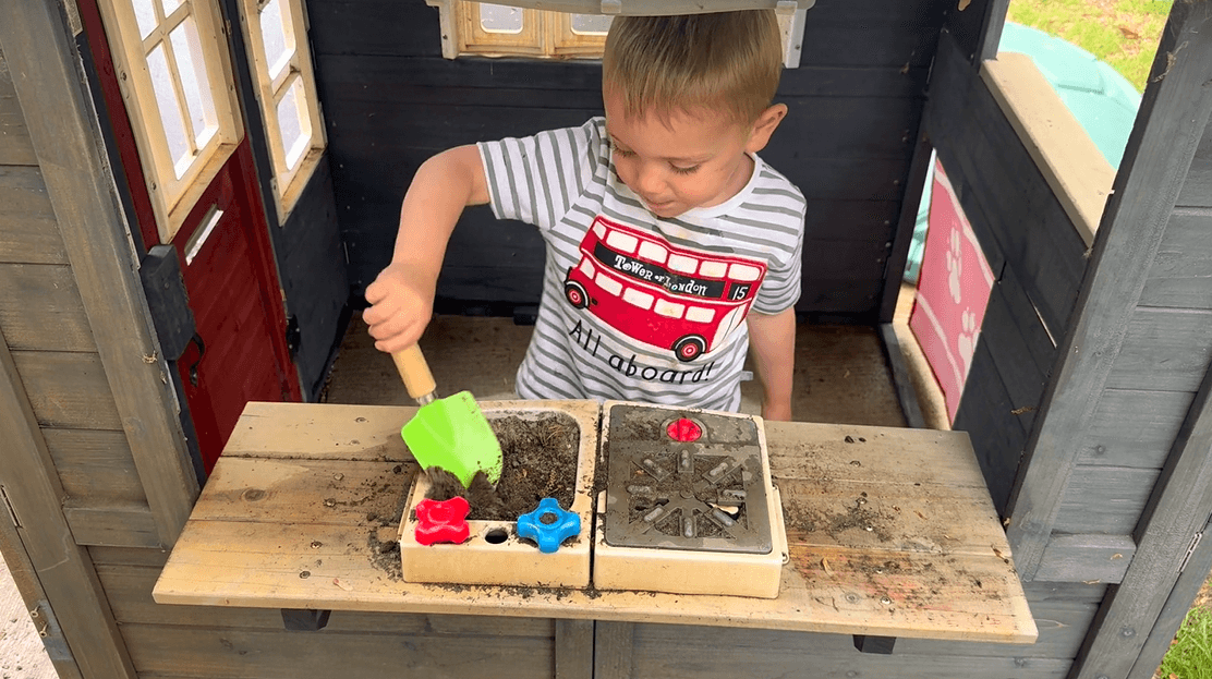 Young boy mixing dirt with green shovel in the sink of his play house mud kitchen.