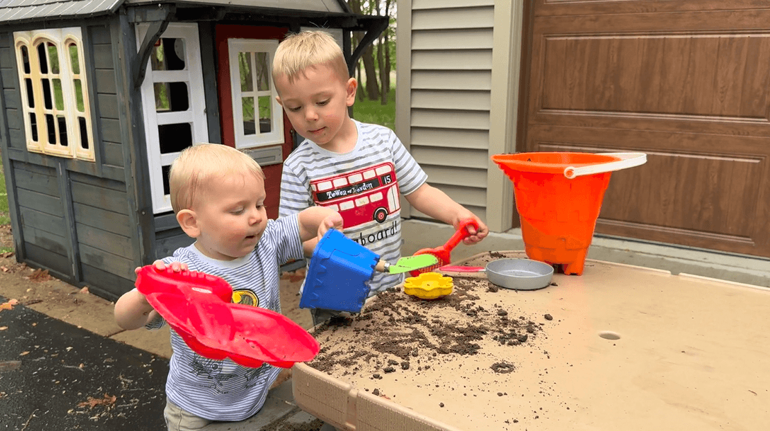 Two little brothers play with dirt on a picnic table that has different mud kitchen tools tools scattered on top.