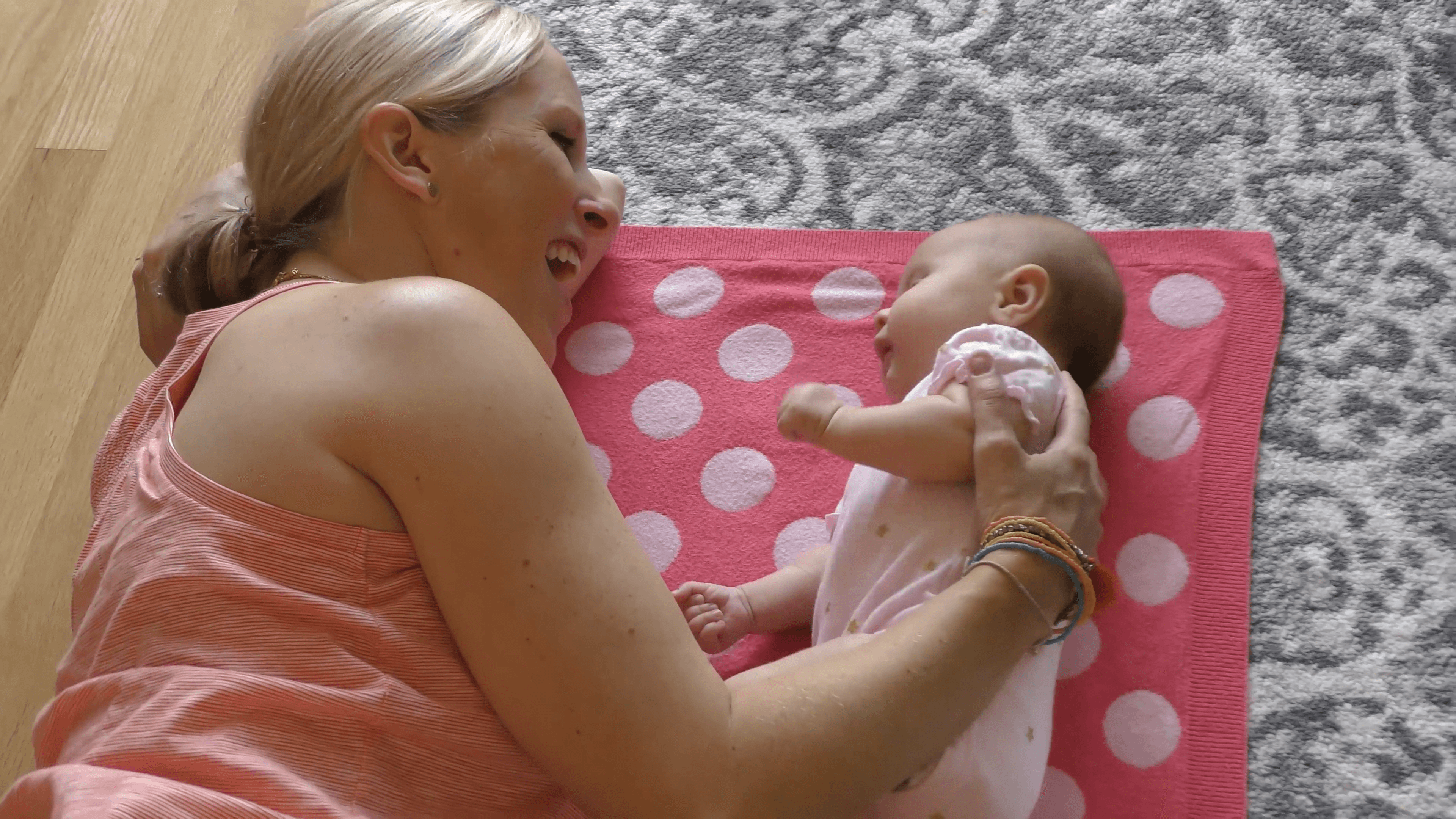 Mother and infant girl lying face-to-face on their sides on top of a pink polka dot blanket. The mother cheerfully engages with her baby.