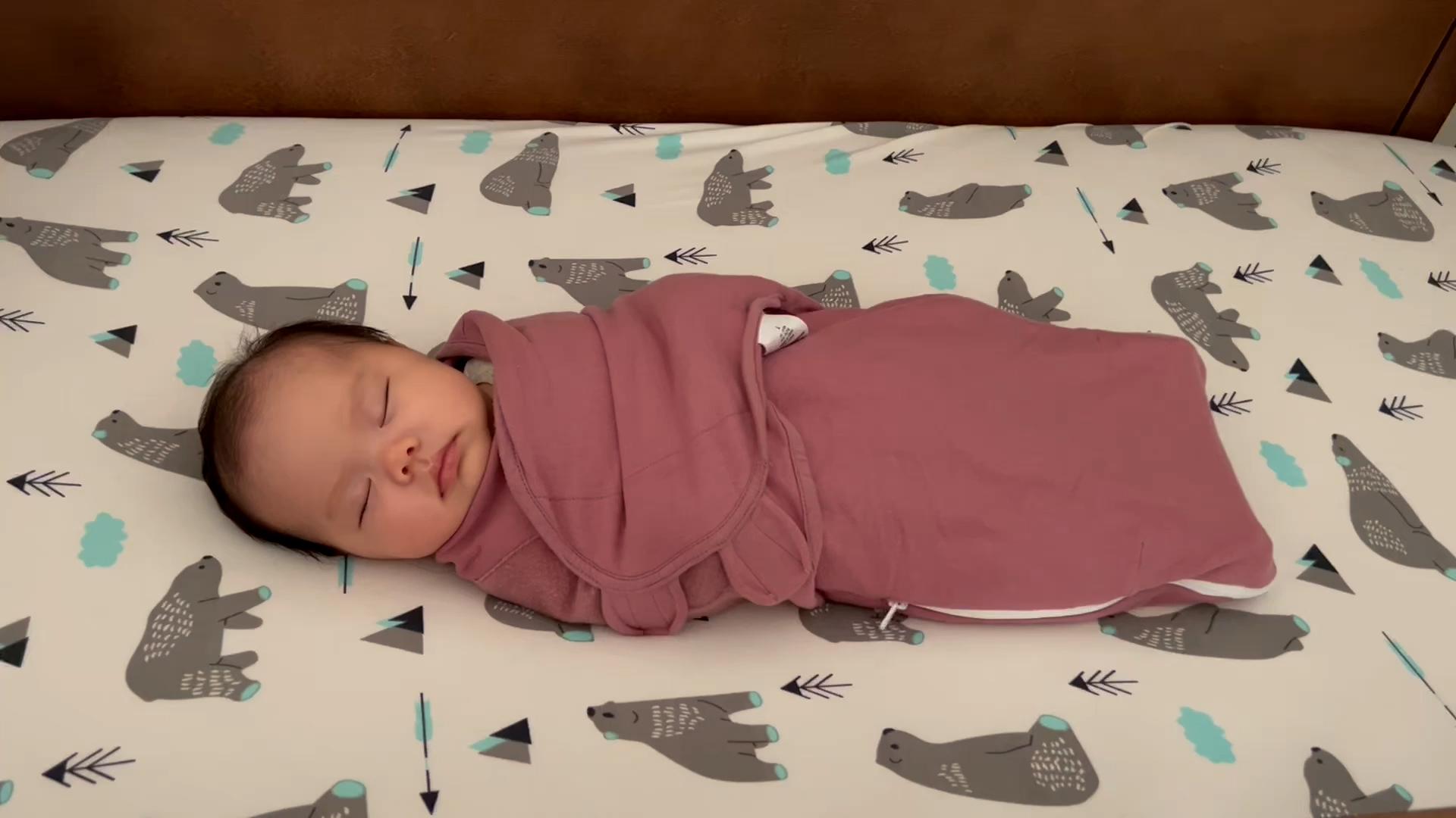 Baby girl sleeping in a mauve-colored swaddle in her crib. The fitted sheet on her crib mattress has a gray-and-blue polar bear pattern all over it.