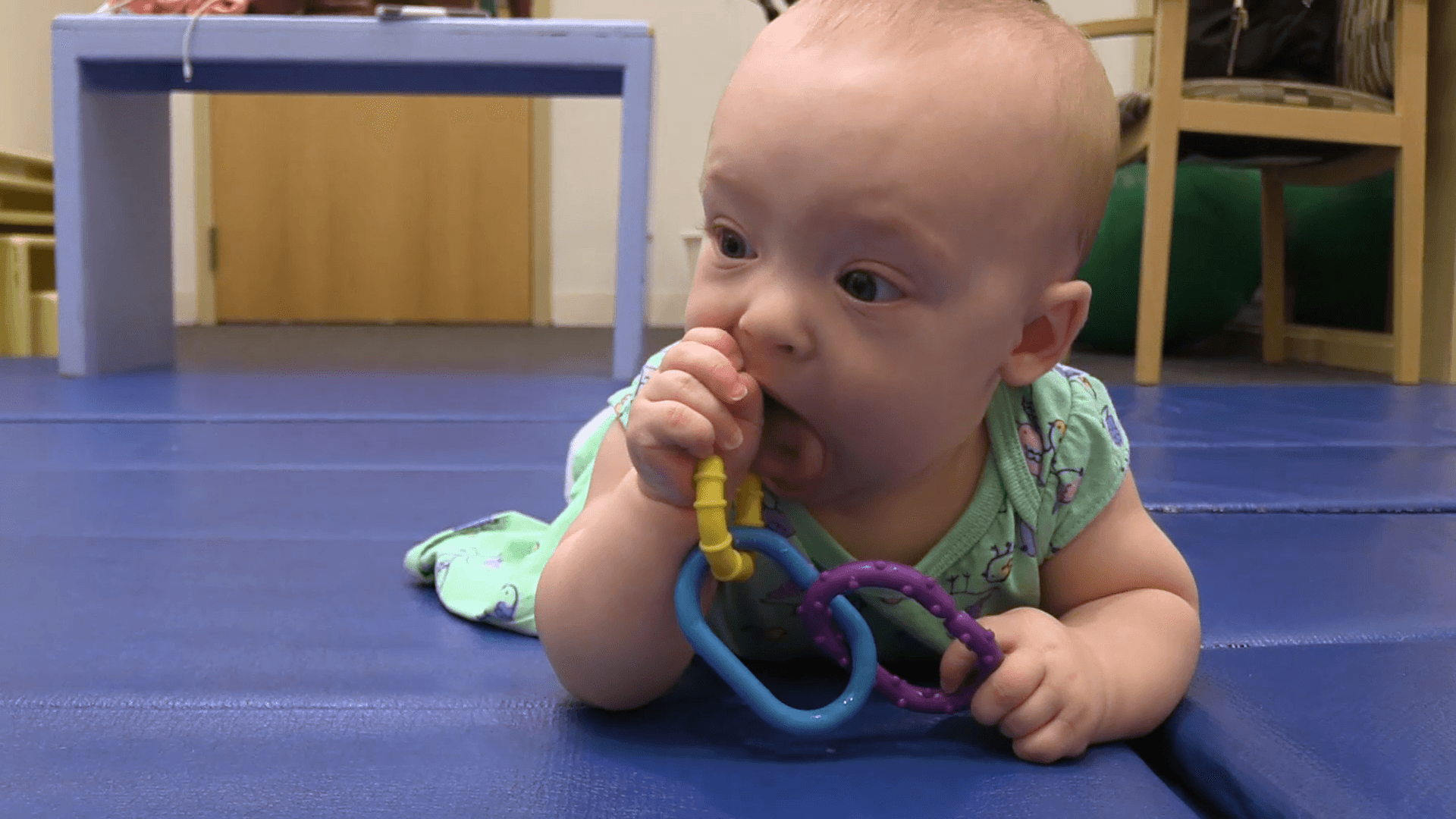 Baby girl doing Tummy Time on blue mat while holding linked rings teething toy in her mouth.