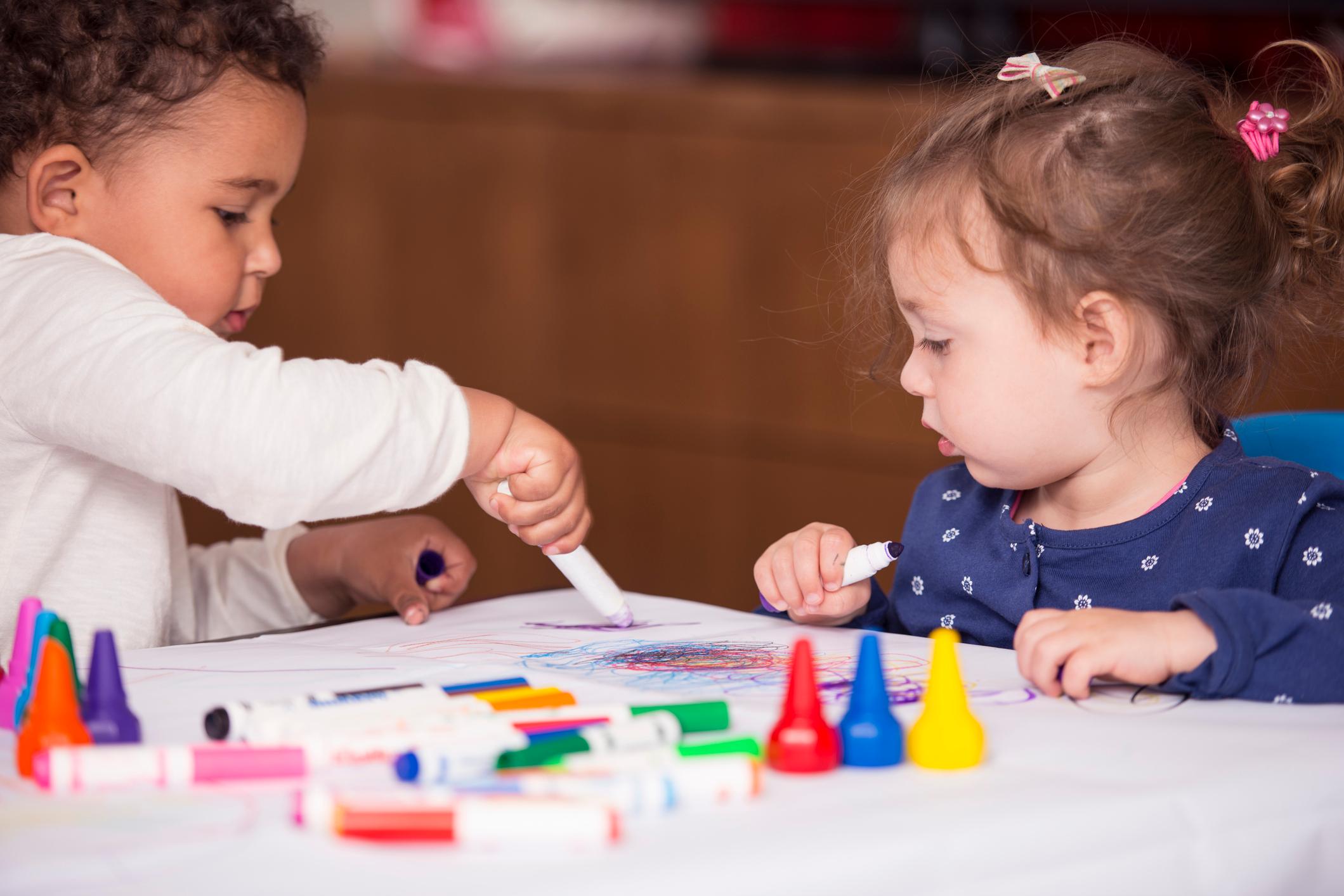 Two toddlers scribble with markers on a piece of paper. Jumbo markers and finger crayons are scattered across the table.