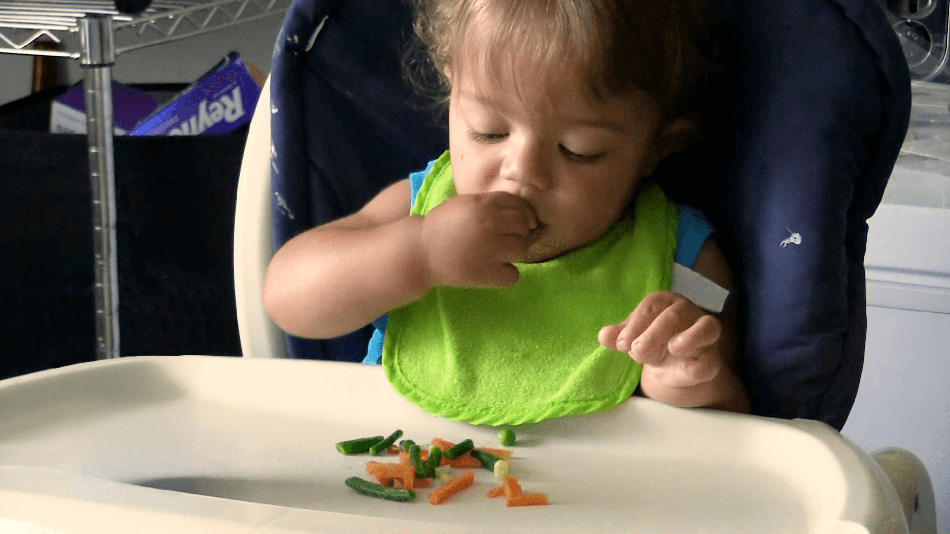 Baby boy with green bib picking up and eating mixed veggies from his high chair tray.