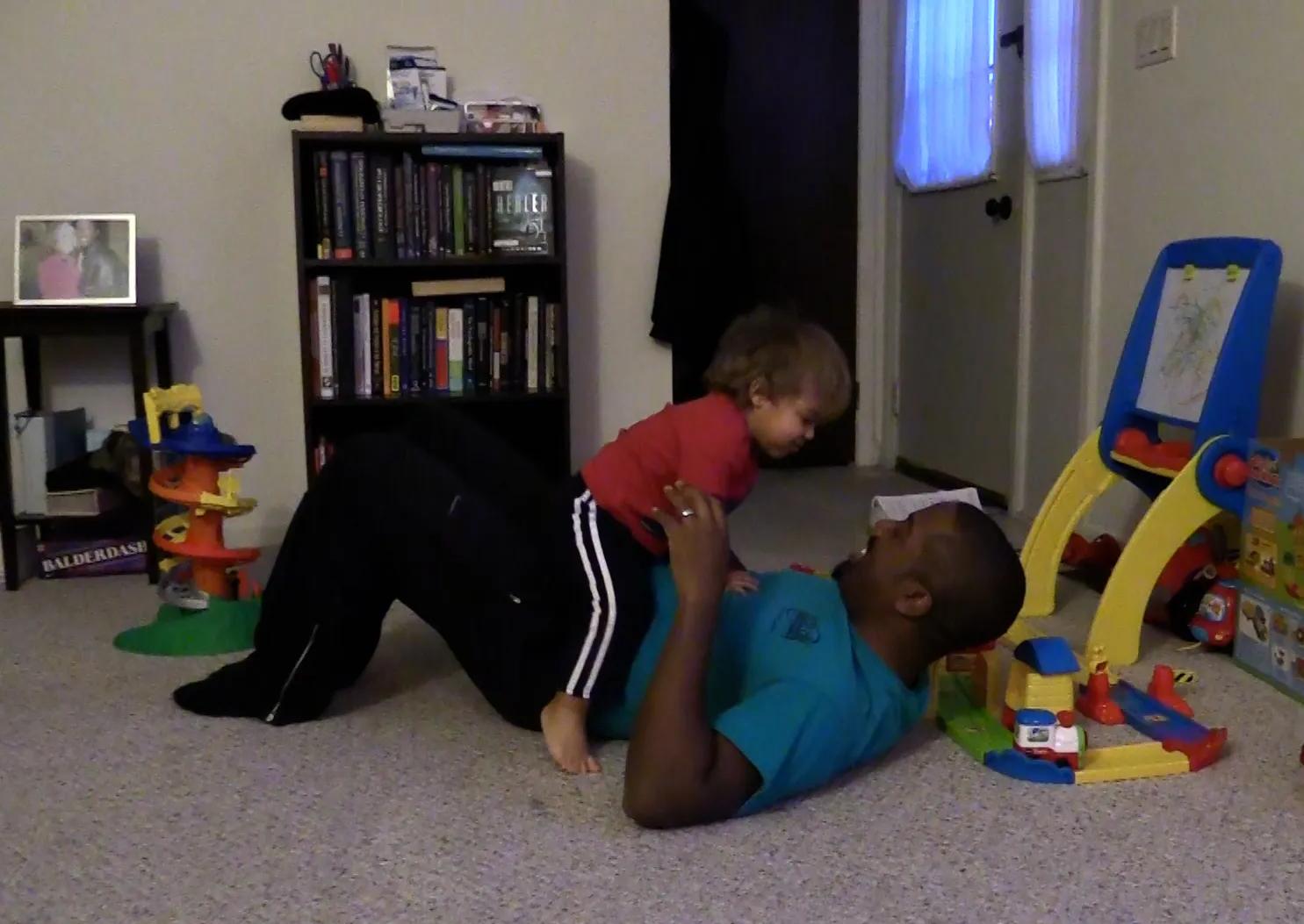 Smiling father holds toddler while lying on his back, surrounded by kids’ toys.