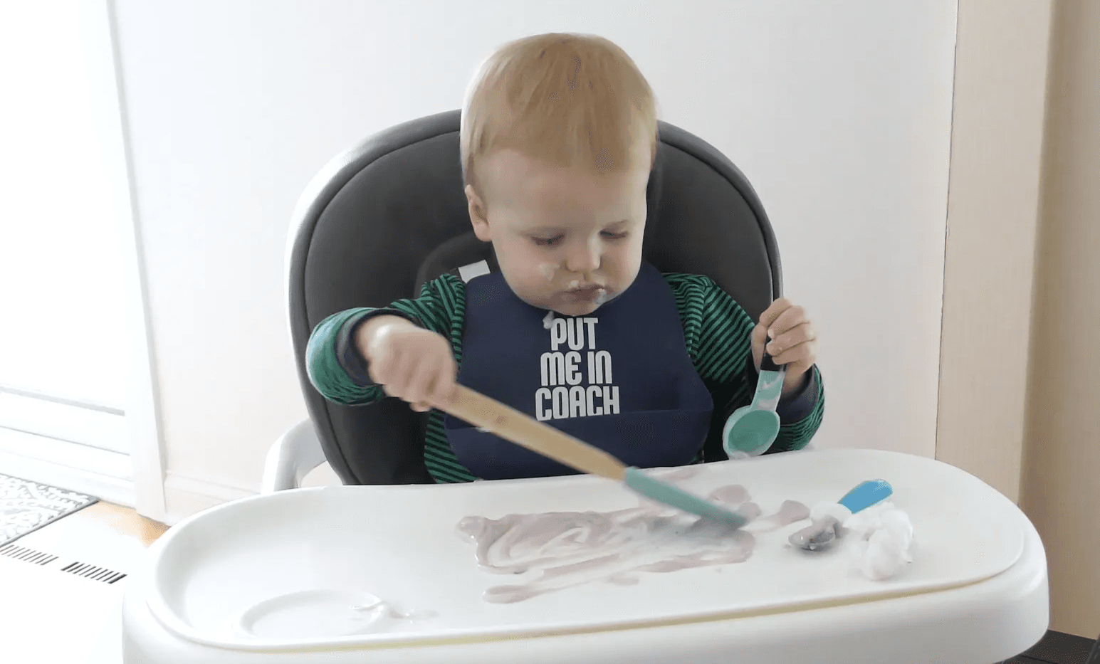 Toddler boy sitting in high chair using a spatula and measuring spoon to play with purple yogurt on his tray.