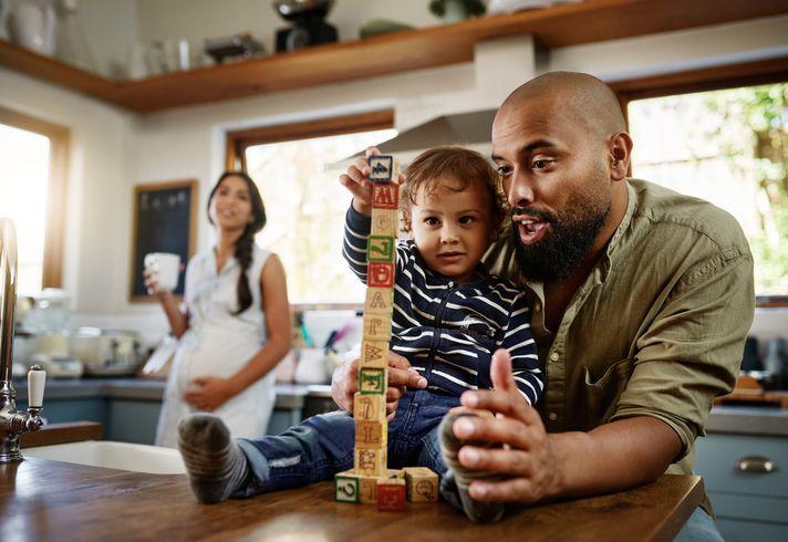 Father and toddler building a tall tower with wooden alphabet blocks on a kitchen counter, while a pregnant mom smiles in the background holding a mug and her belly.