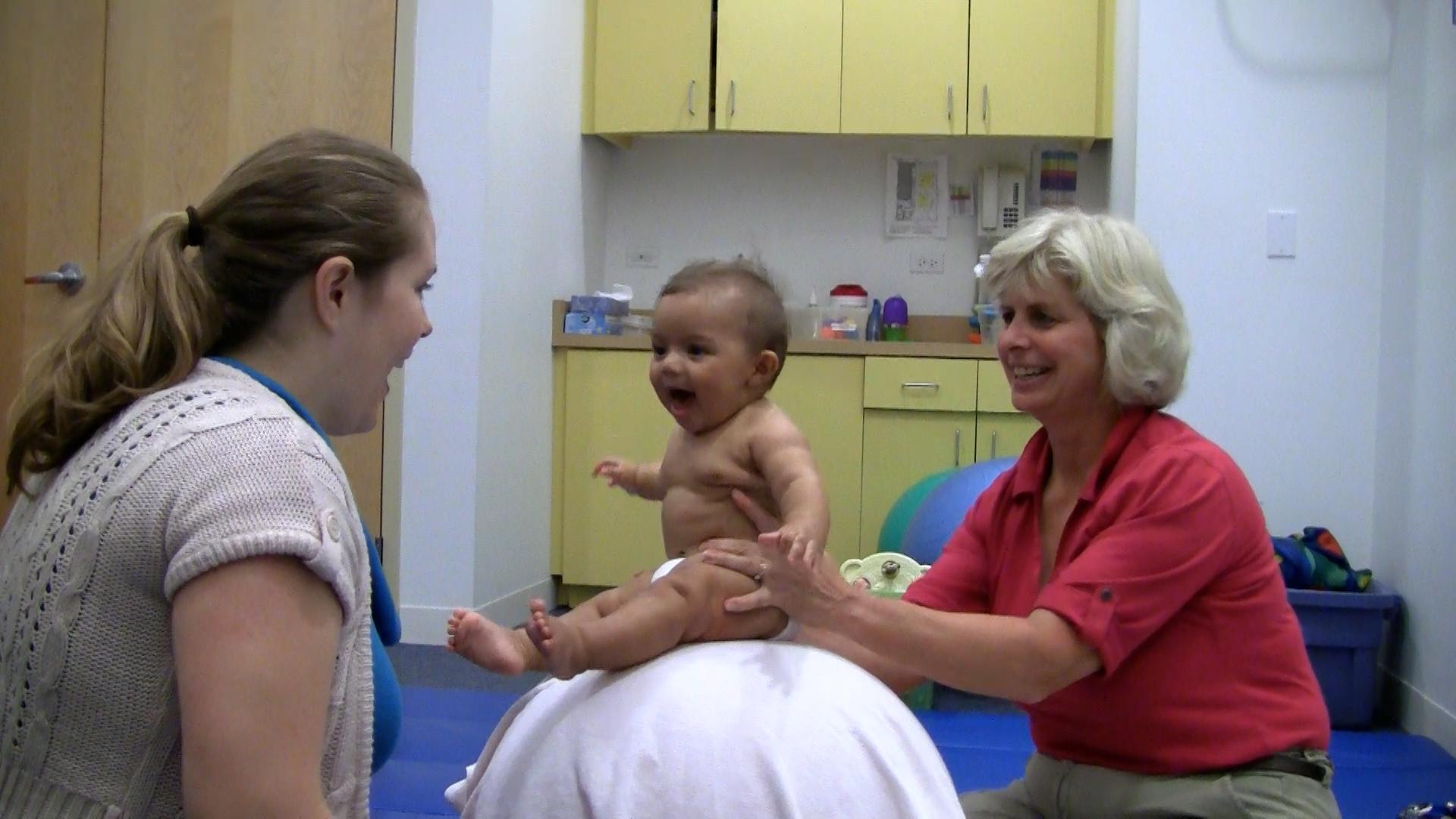 Pediatric therapist holds baby boy on an exercise ball as he babbles happily at his mother.