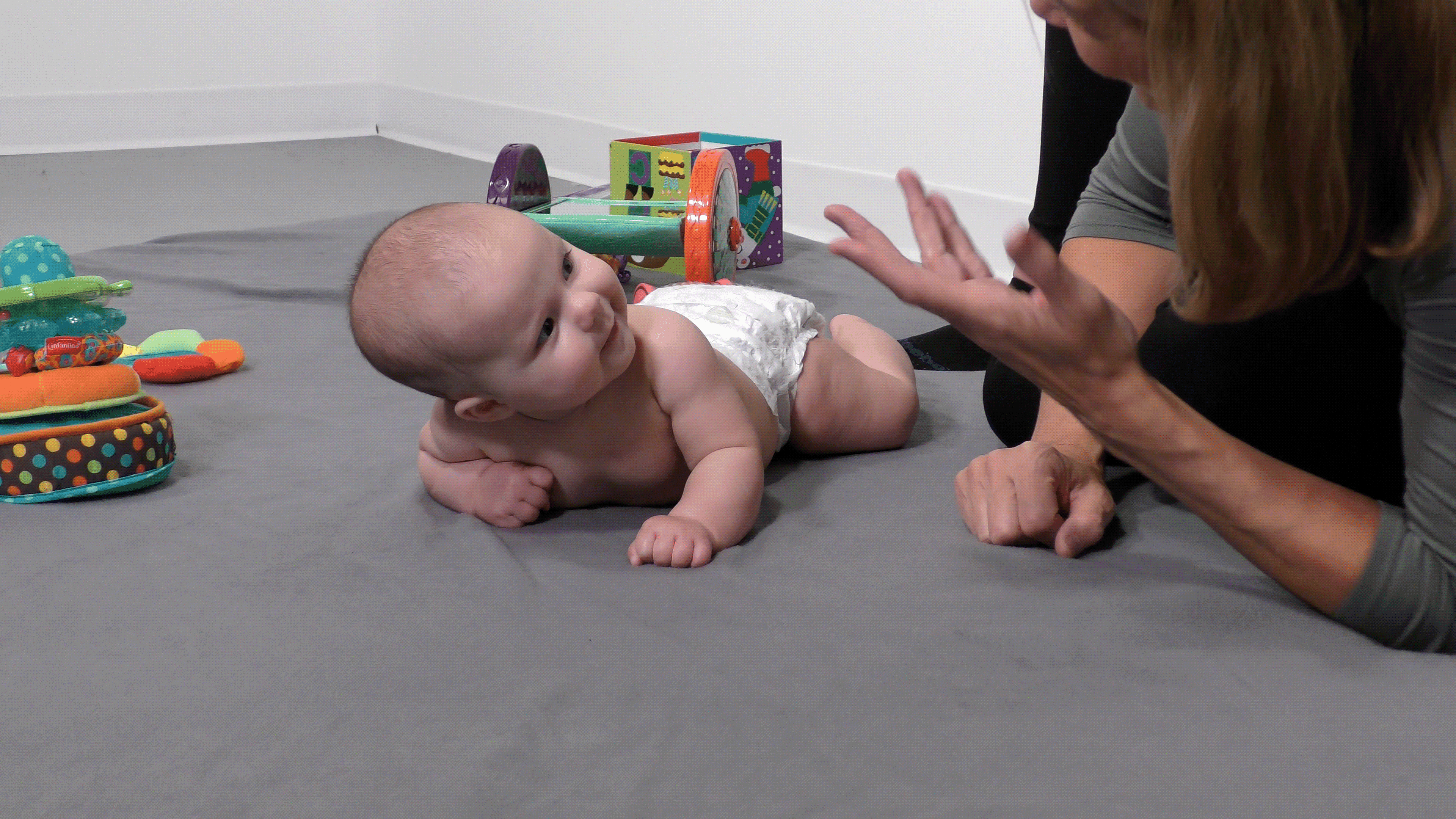 Baby lying on tummy and tilting its head to look at a nearby caregiver, who has her hand out as if she’s asking “why?”.
