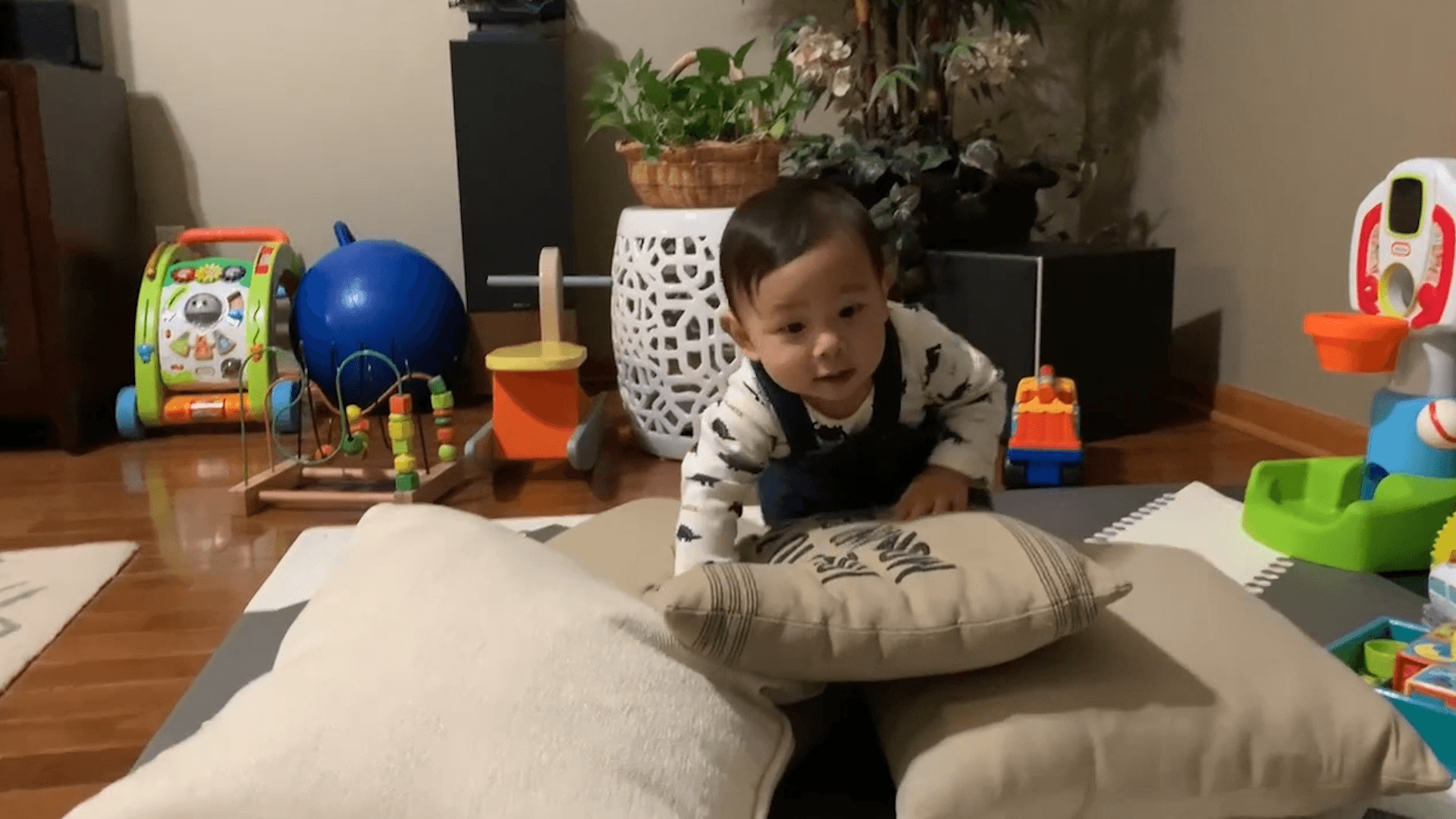 Older baby boy crawling on top of pillows on the living room floor. A collection of big toys like a mini basketball hoop and an activity walker can be seen in the background.