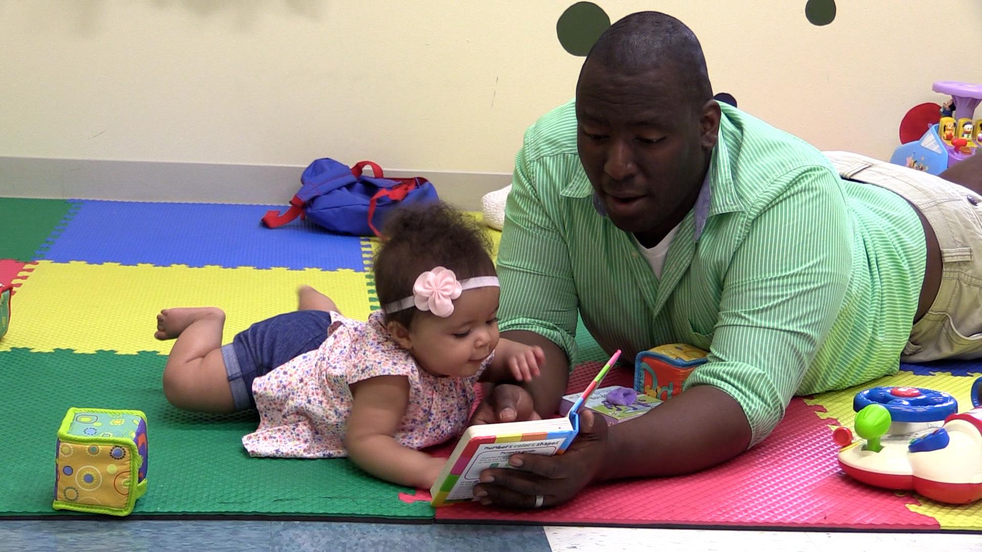An older baby girl and her dad are reading a book while lying on their stomachs. They are lying on top of a colorful foam puzzle piece floor with baby toys scattered about.