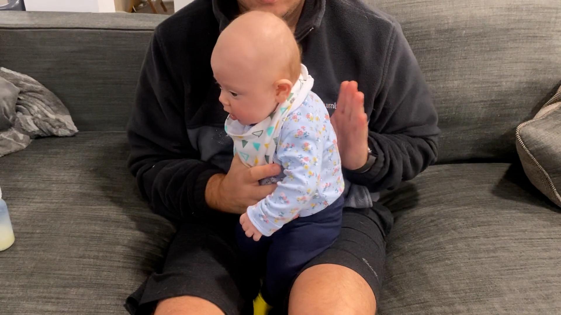 A dad sits on a gray couch while burping his baby boy, who is held upright on his lap.