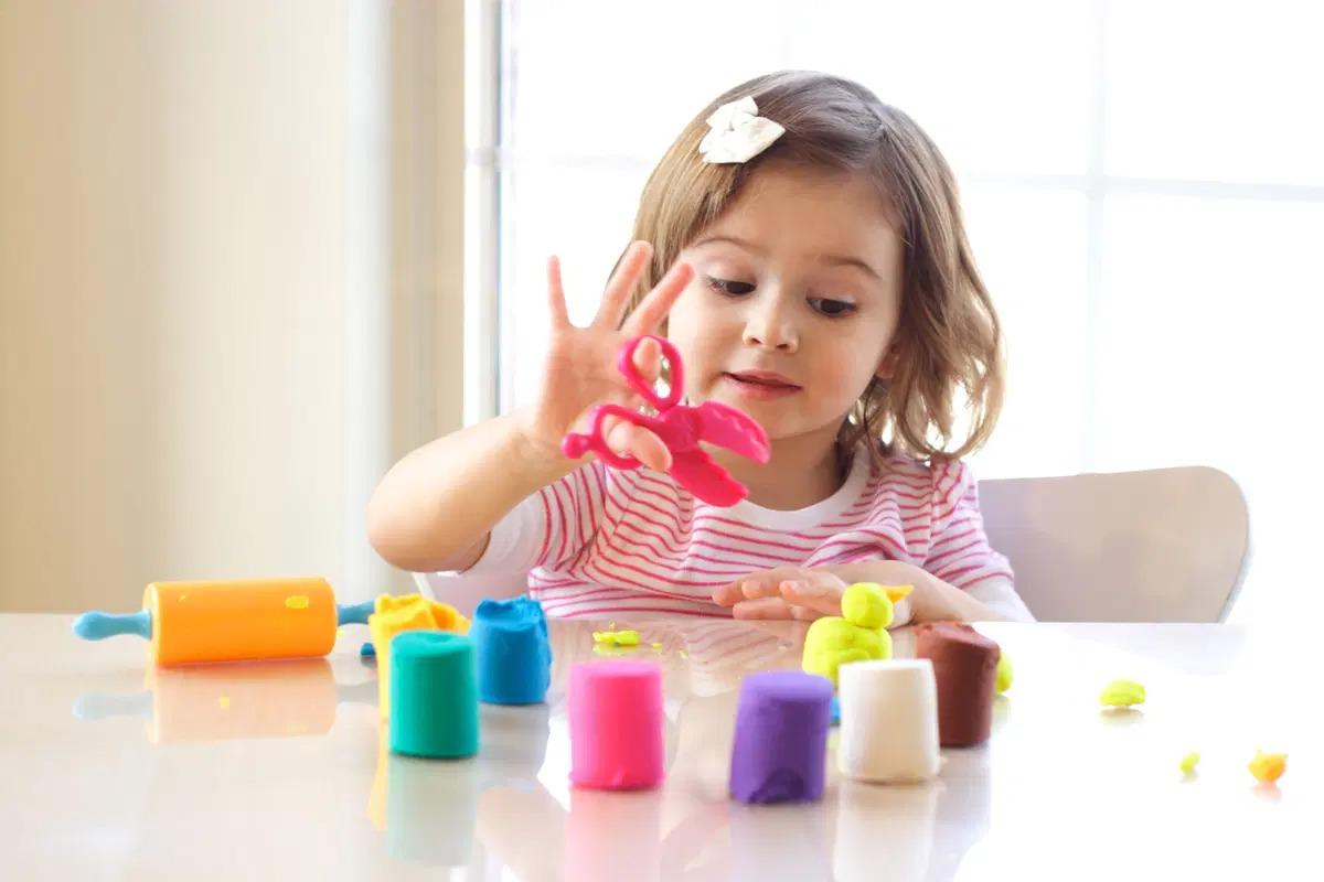 Young child using plastic scissors to cut colorful play-dough at a table.