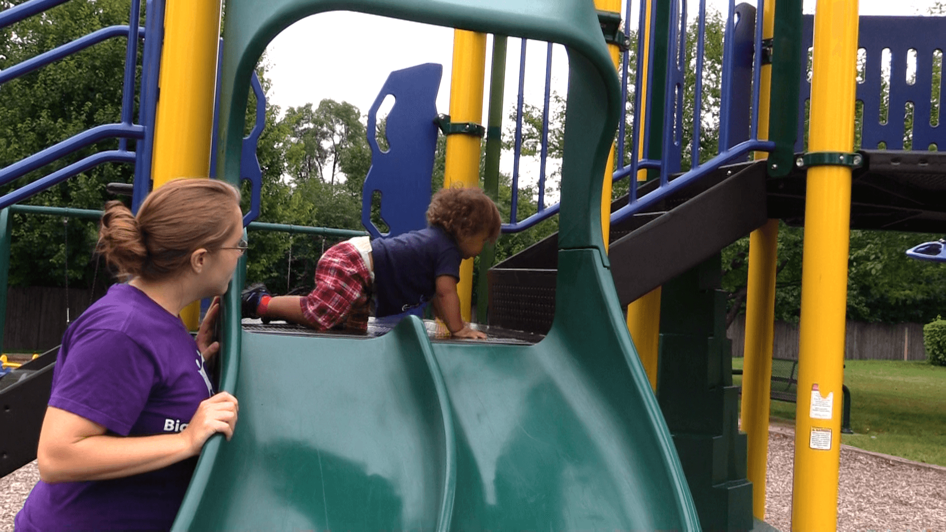 Toddler crawling on a playground structure as his mother watches nearby.
