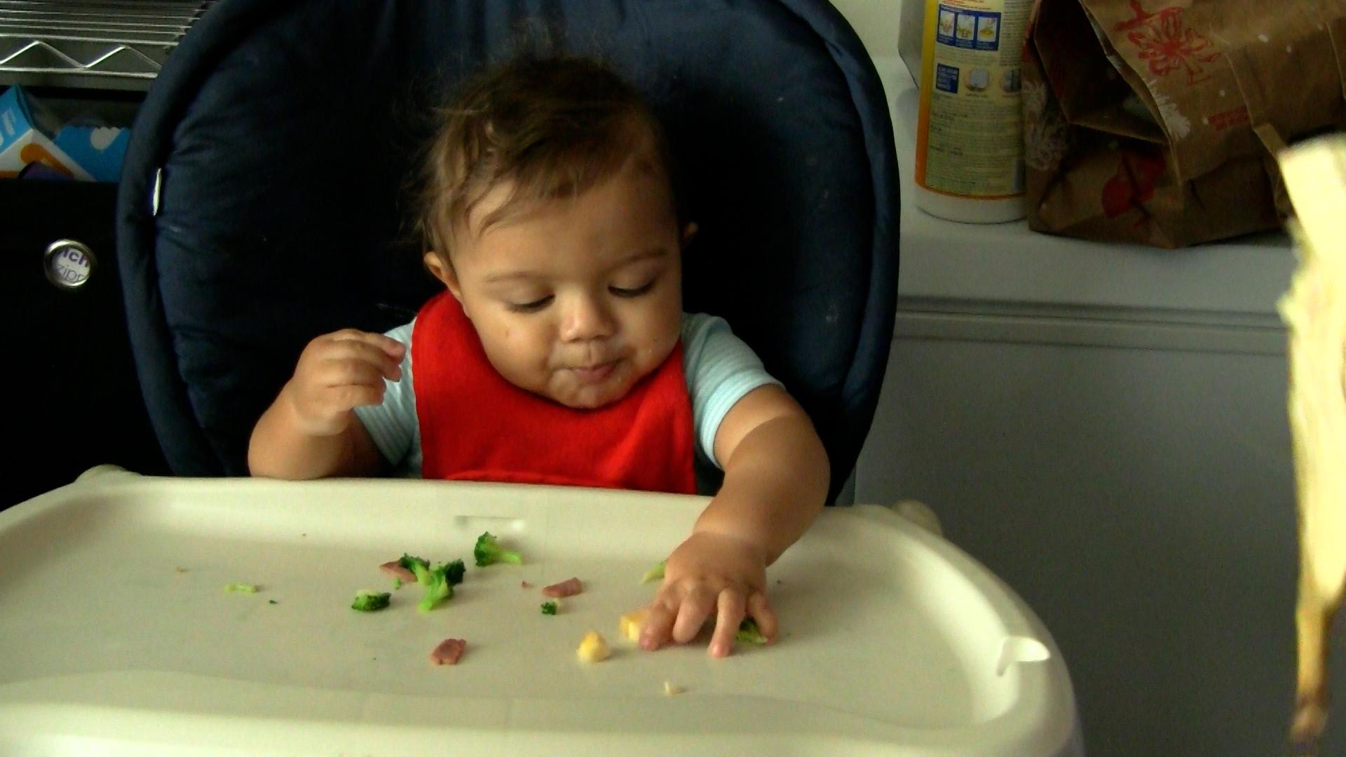Older baby boy sits in his high chair and finger-feeds himself small pieces of broccoli, ham, and corn.