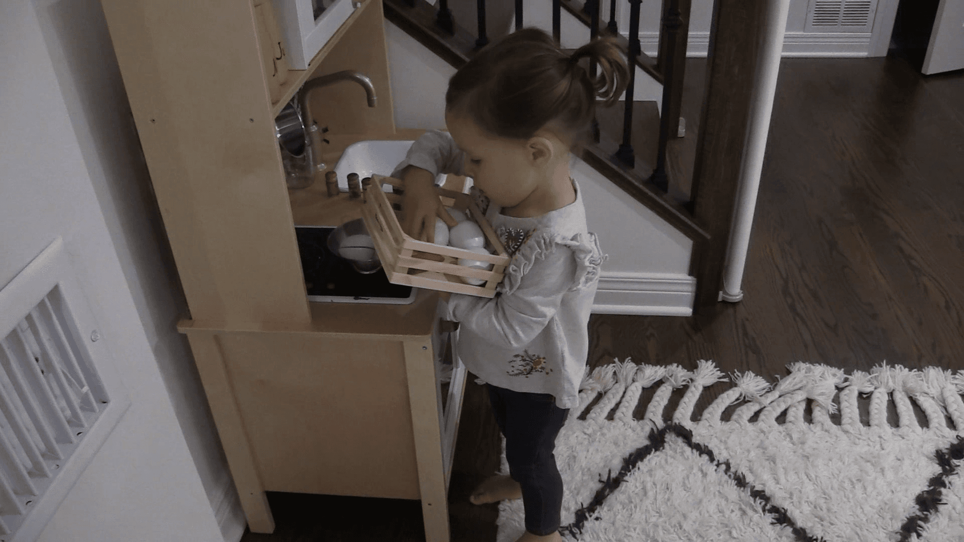 Toddler girl collecting plastic eggs while standing at a wooden play kitchen.