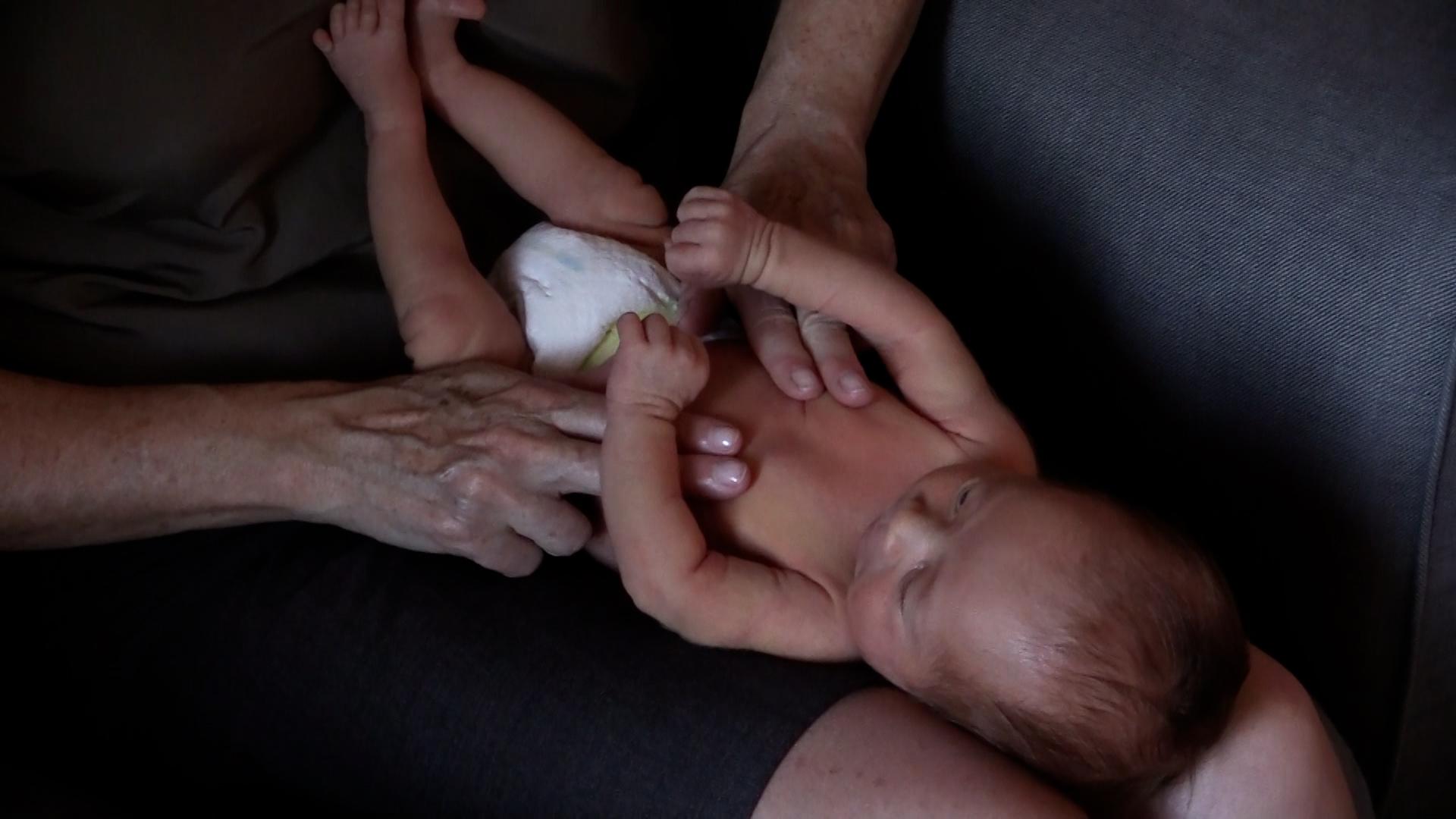 A newborn baby receives a baby massage while lying across a woman’s lap. The woman uses two fingers on each hand to apply gentle, deep pressure on the newborn’s belly.