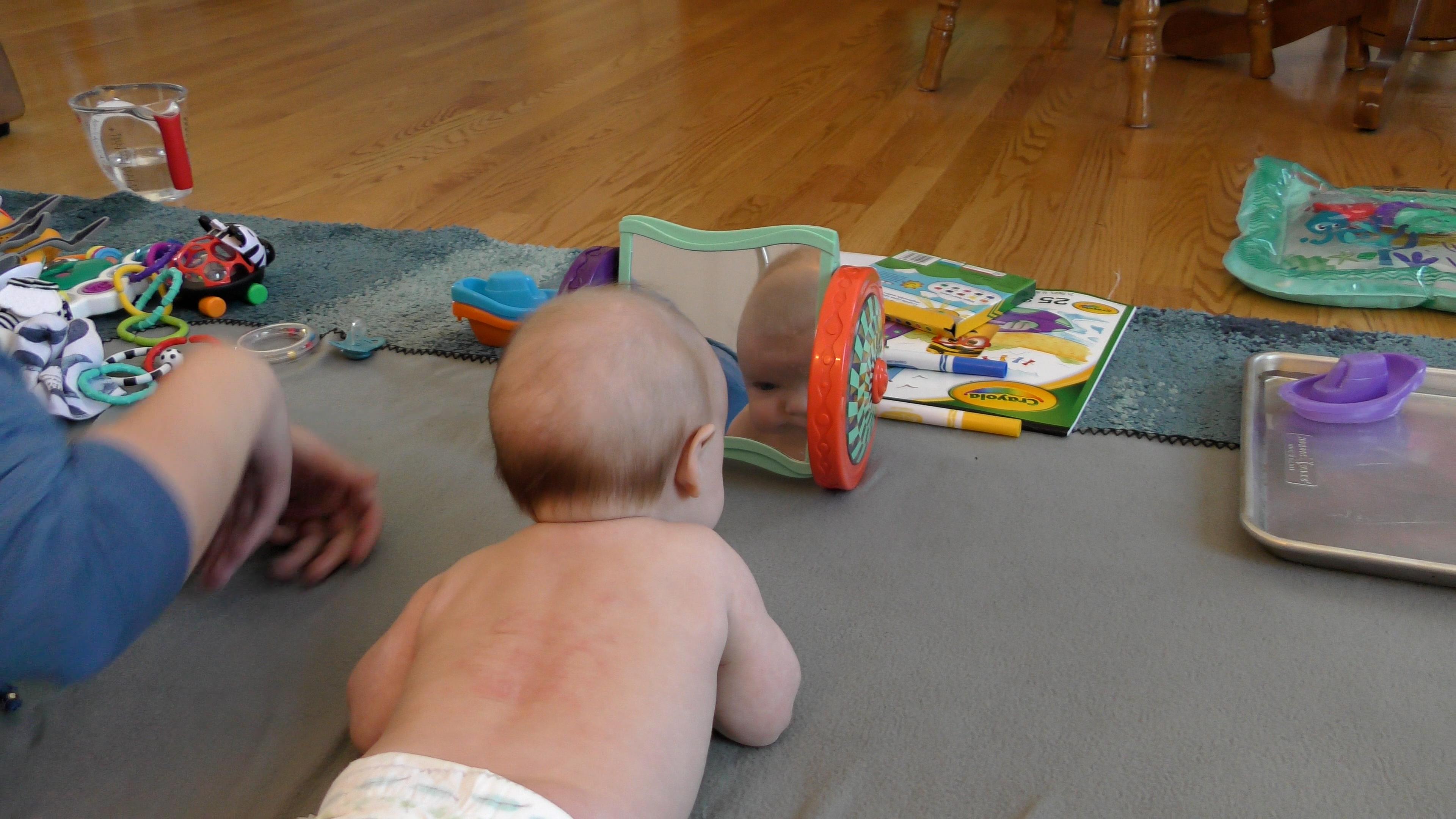 Baby boy looking into baby mirror while doing Tummy Time on a rug that has toys scattered around it.