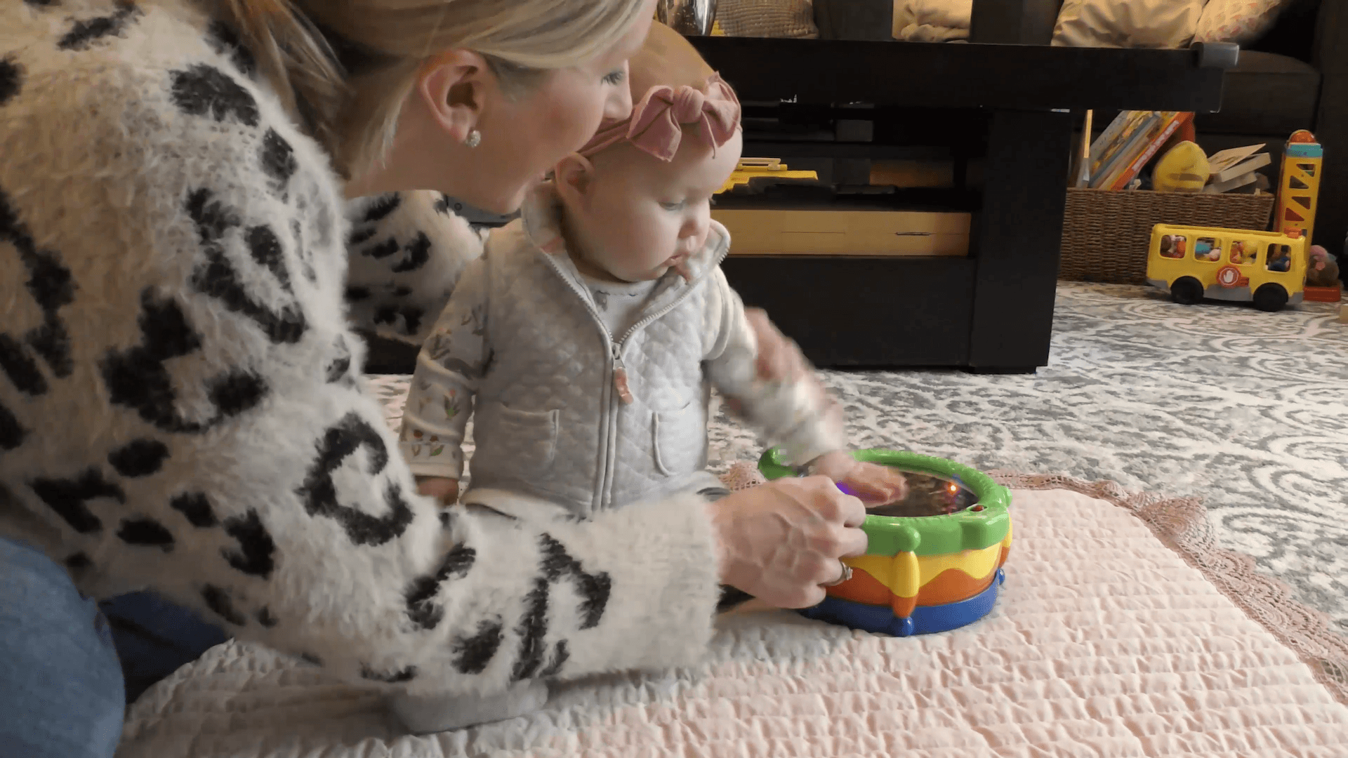 Mom interacting with baby girl sitting and playing with toy drum on the living room floor.