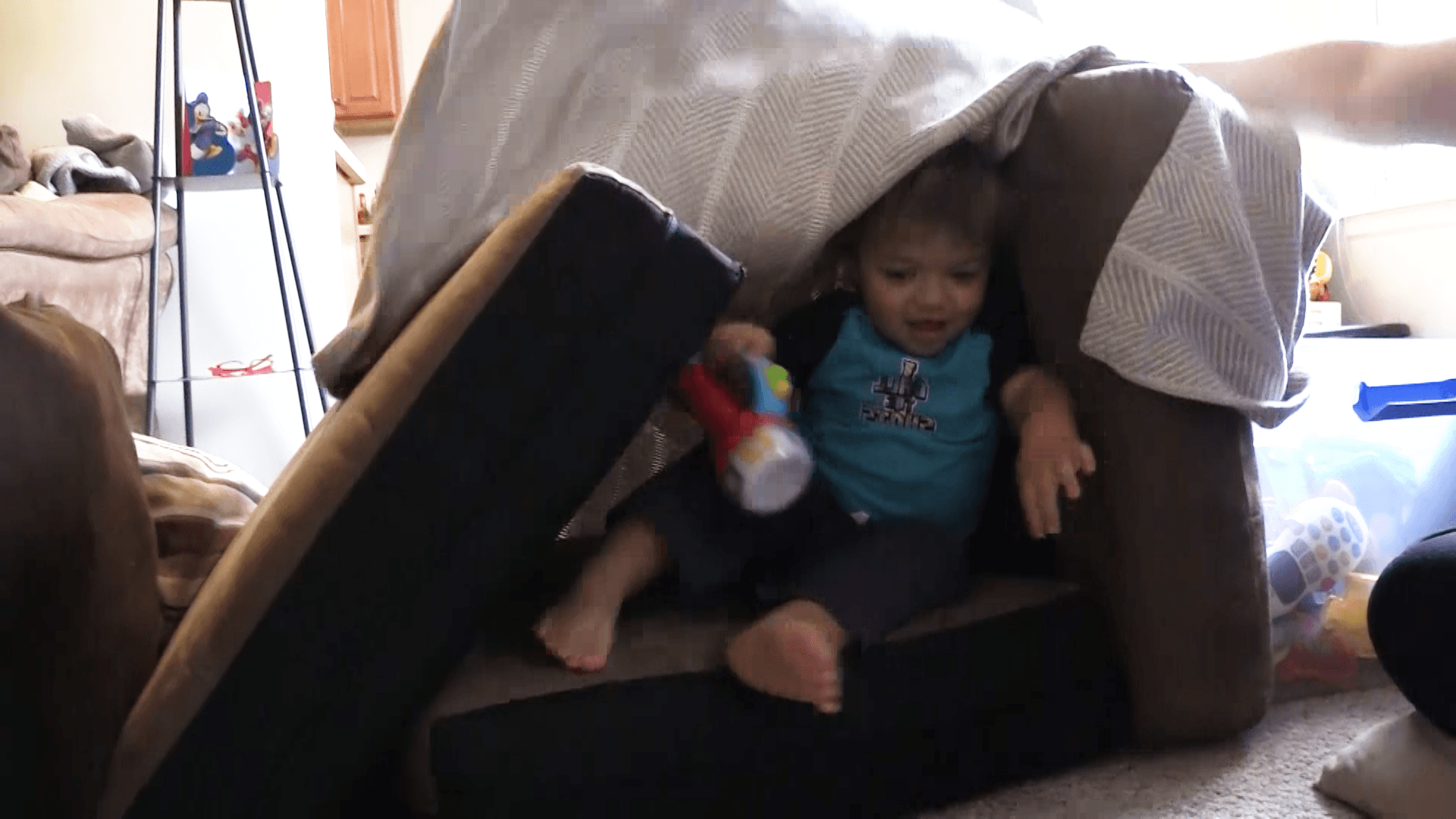 A smiling toddler boy holds a toy flashlight as he emerges from a fort made out of couch cushions.
