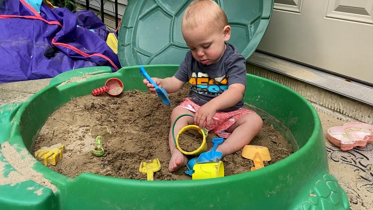 A toddler boy sits in a turtle-shaped sandbox and plays with various sand tools.