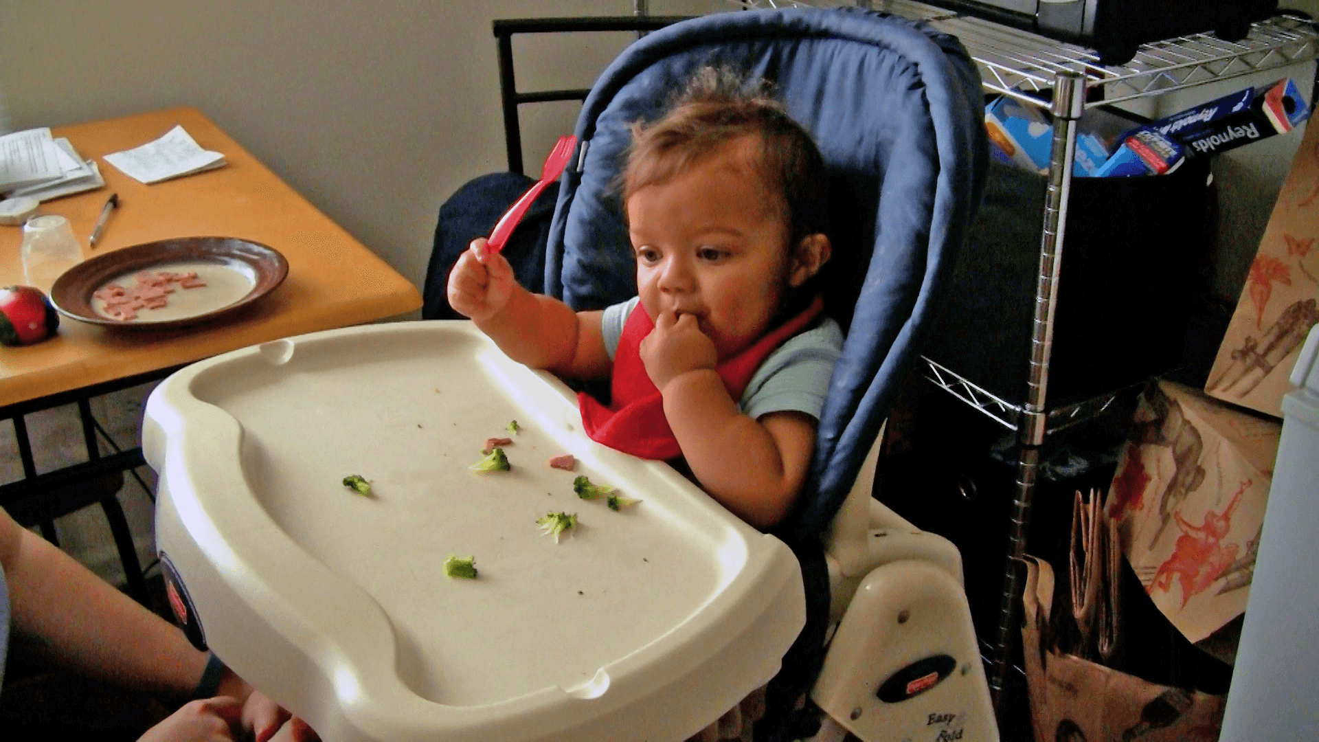 Baby boy feeding himself small broccoli pieces from his high chair tray while holding a red spoon in the other hand.