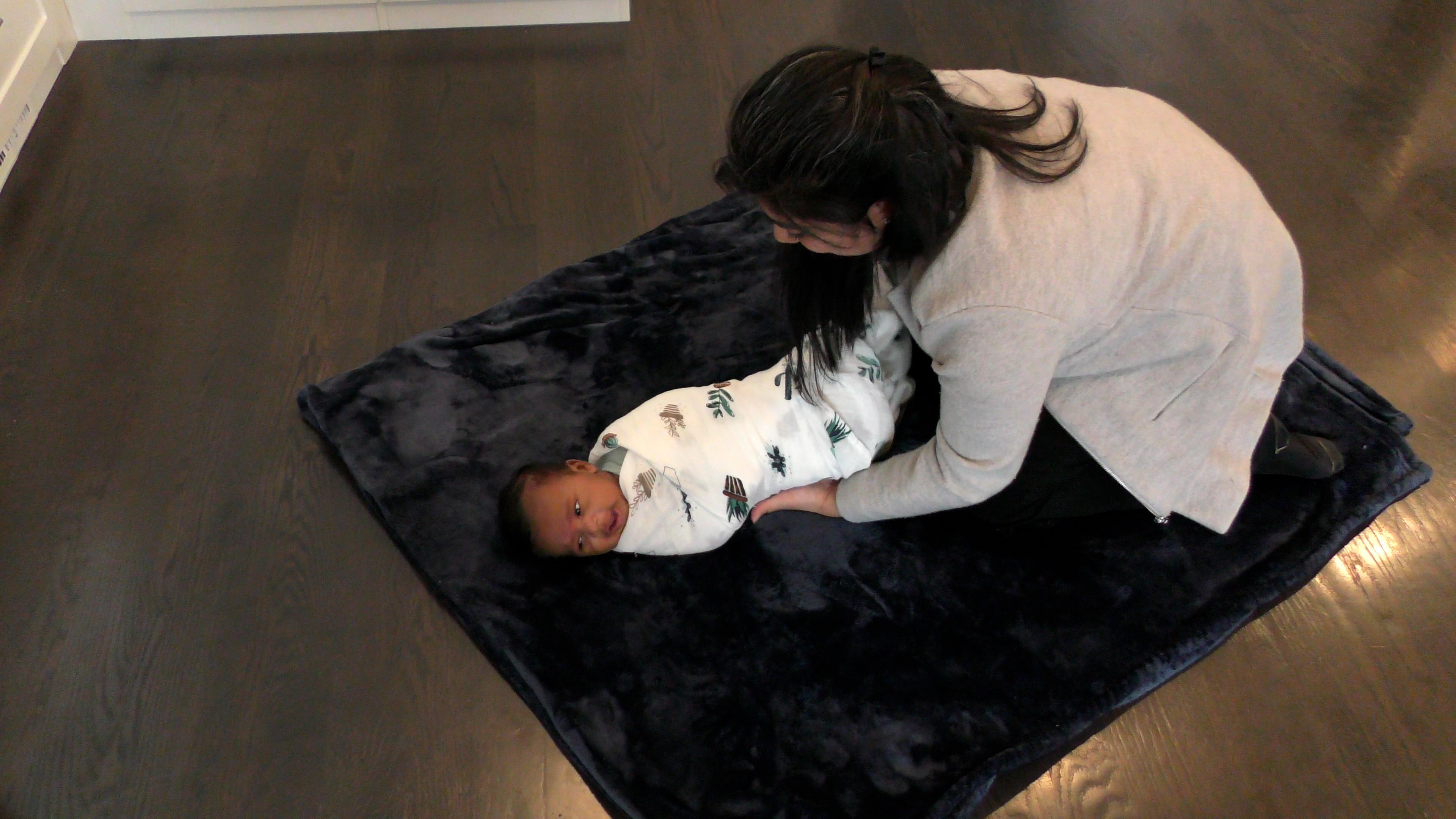 Woman in a gray jacket swaddles a baby boy while she kneels over him on a soft navy blanket.